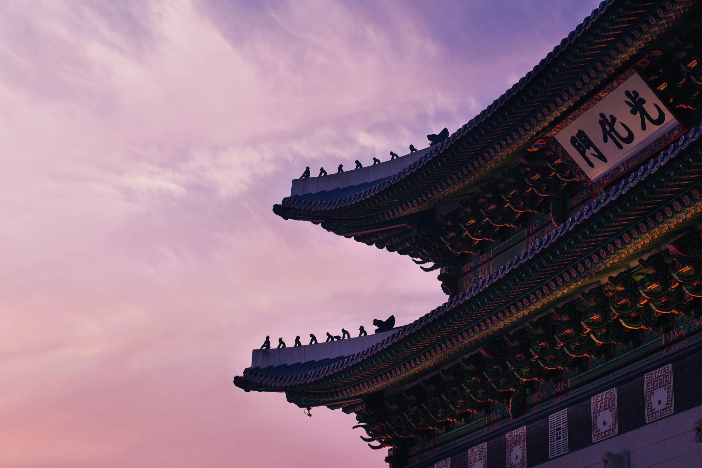 Silhouette of traditional Asian architecture against a pink and purple sunset sky, featuring ornate roof details and decorative figures.