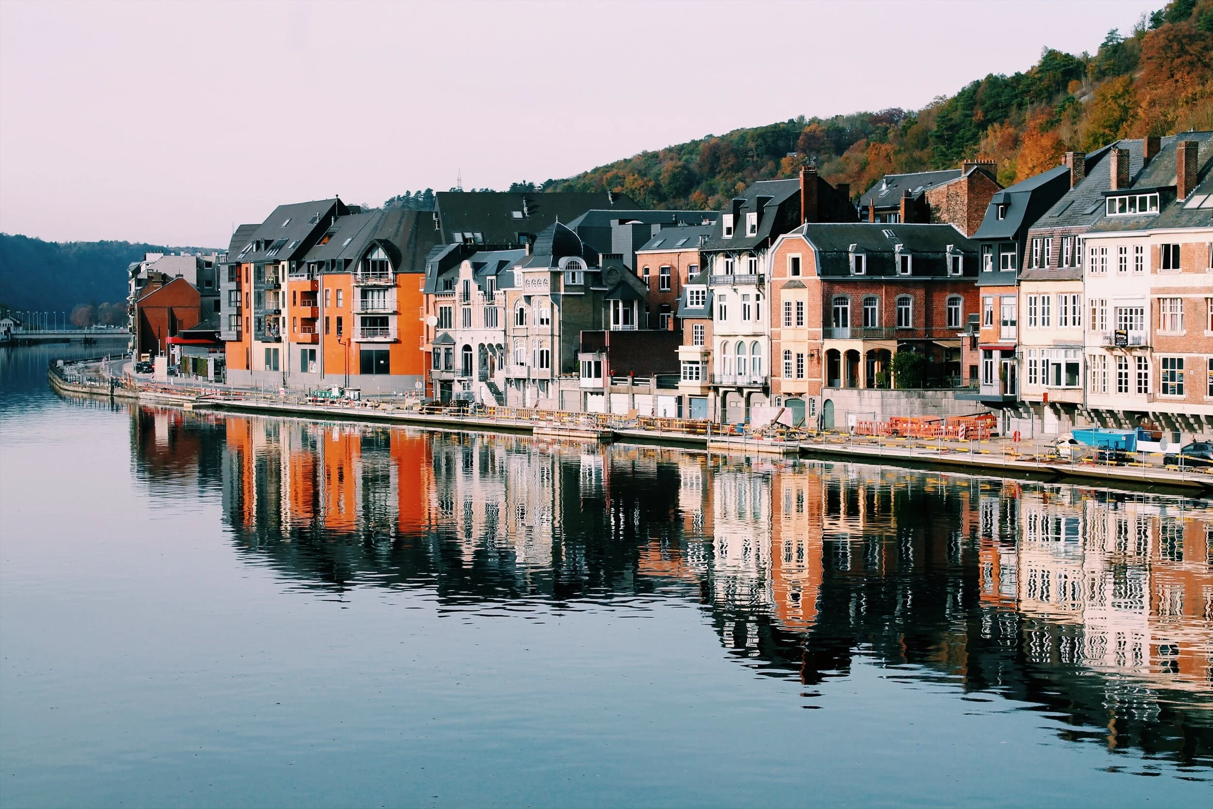 Colorful waterfront buildings reflected in calm river water, with hills in the background.