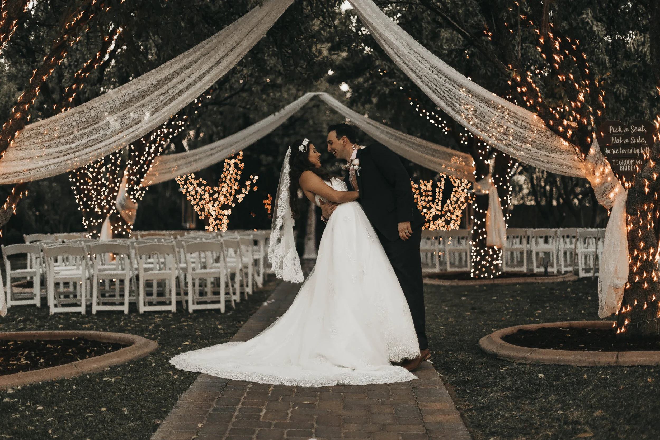 A bride and groom embracing under a decorated canopy with string lights in an outdoor wedding ceremony setting, featuring white chairs and draped fabrics.