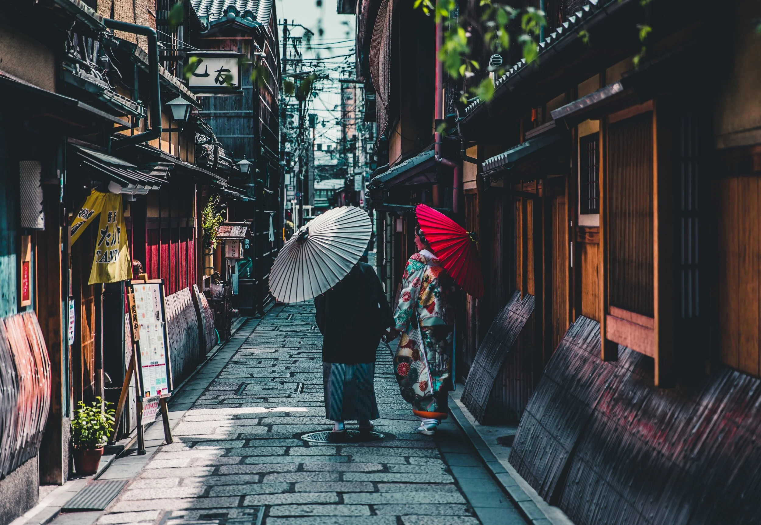 Two people walking down a traditional Japanese street, wearing kimonos and holding parasols. The street is lined with wooden buildings and signs.