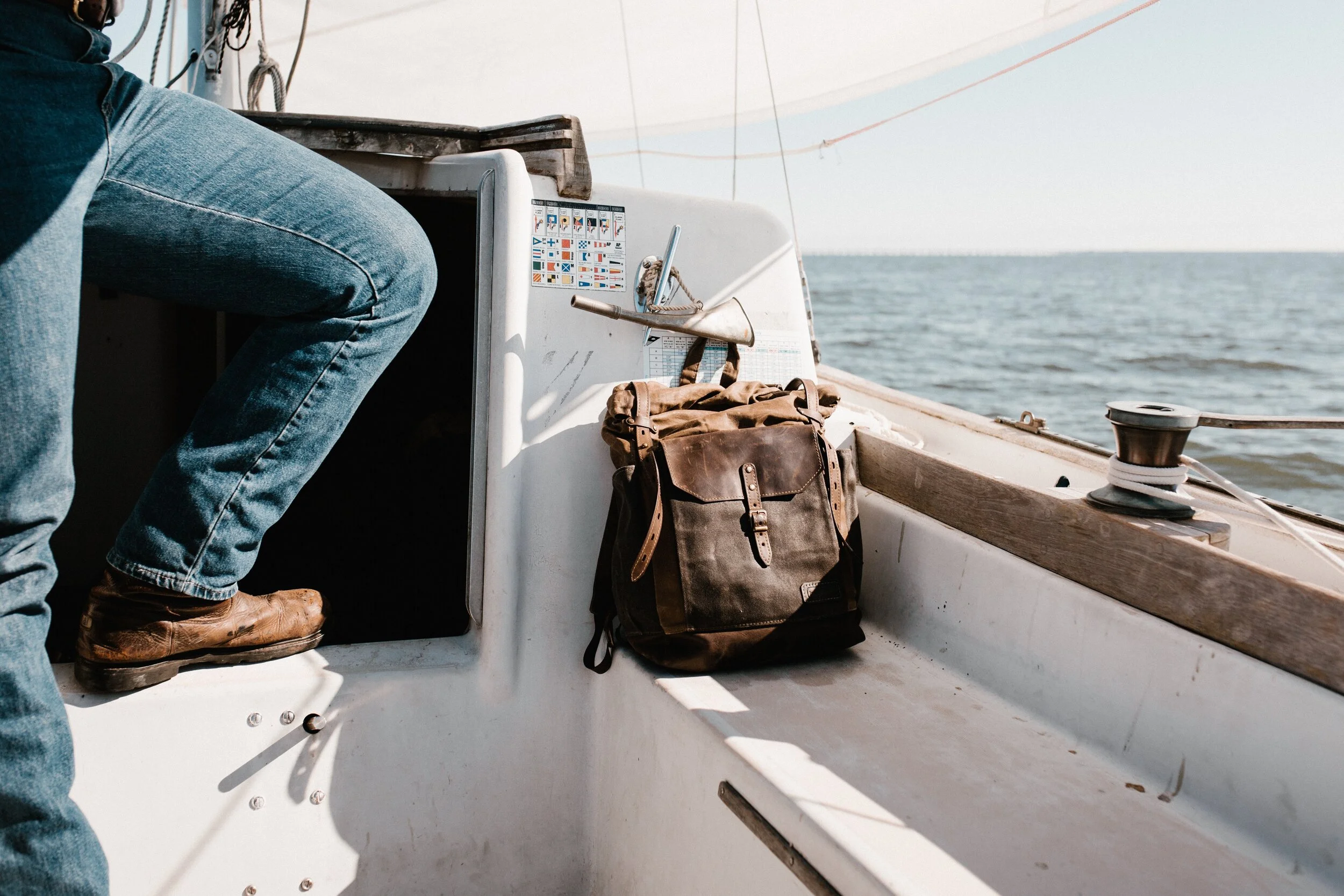 Man in jeans on a boat with a brown backpack on a bench, sea in the background.