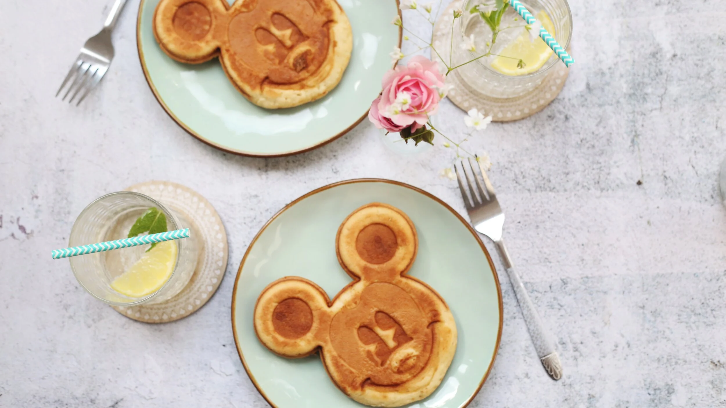 Plates with Mickey Mouse-shaped pancakes, two forks, glasses with lemon water, pink flower, pastel background.
