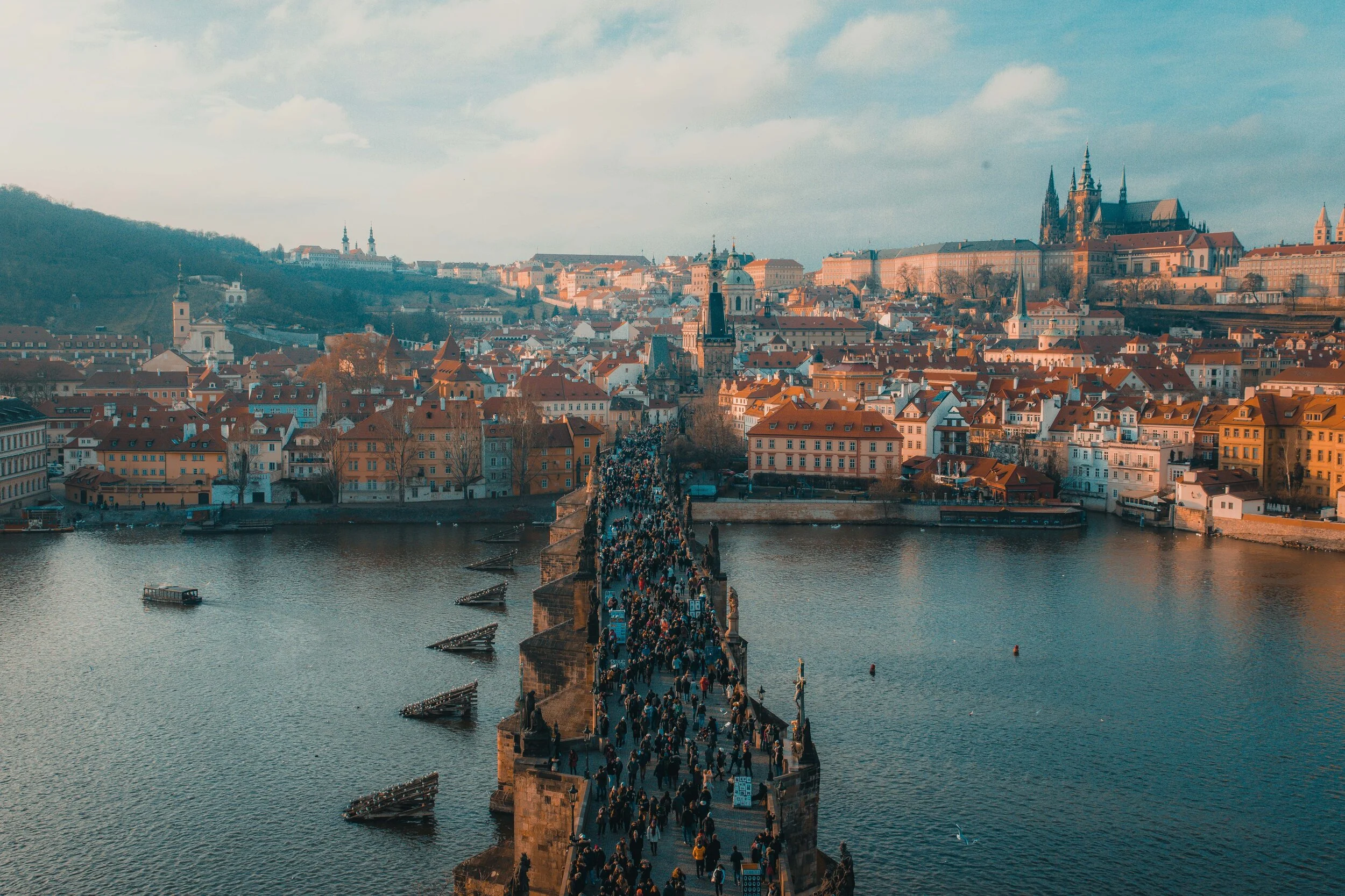 Aerial view of Charles Bridge over the Vltava River in Prague, with a crowd of people walking, surrounded by historic buildings and Prague Castle in the background under a partly cloudy sky.