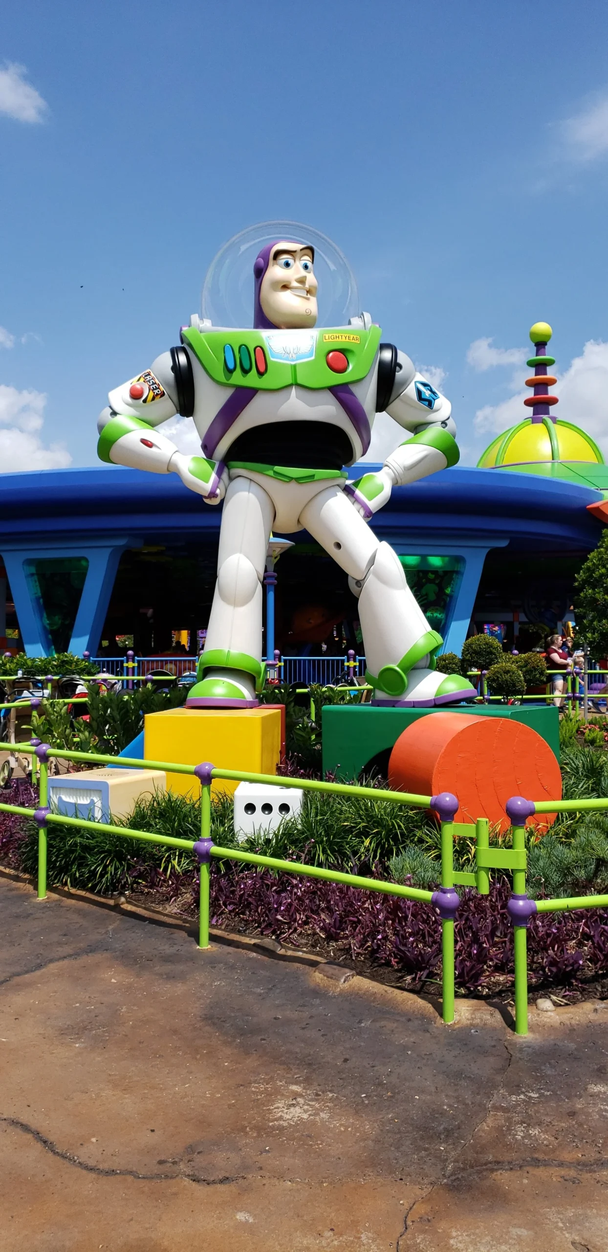Large statue of Buzz Lightyear from Toy Story in an amusement park setting, surrounded by colorful decorations and a blue sky.
