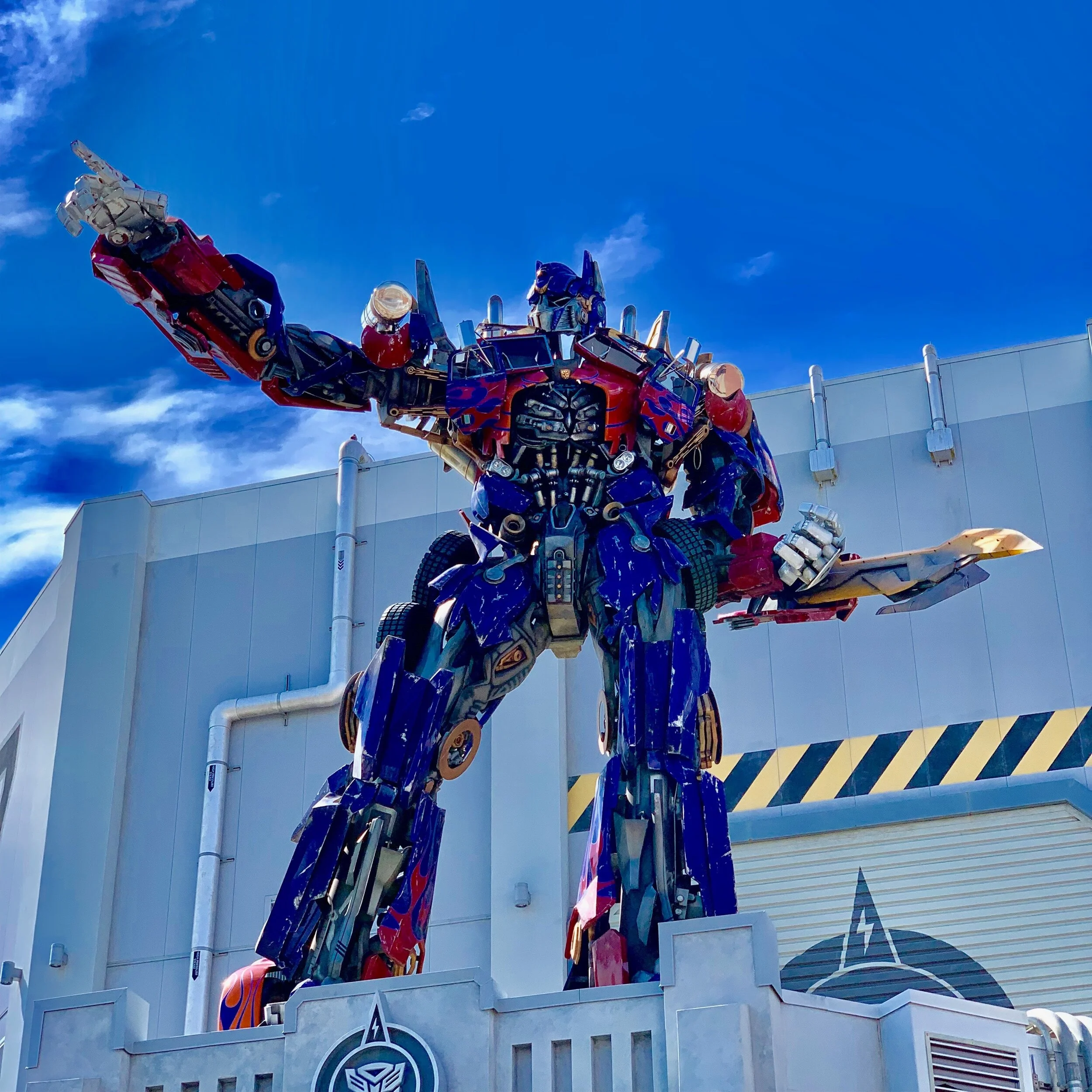 Statue of a large robot resembling a transformer in red and blue colors, positioned in front of a building with a clear blue sky background.