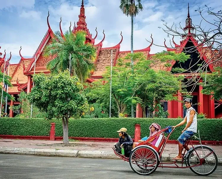 A traditional rickshaw with a driver and two passengers in front of a red ornate building surrounded by greenery.