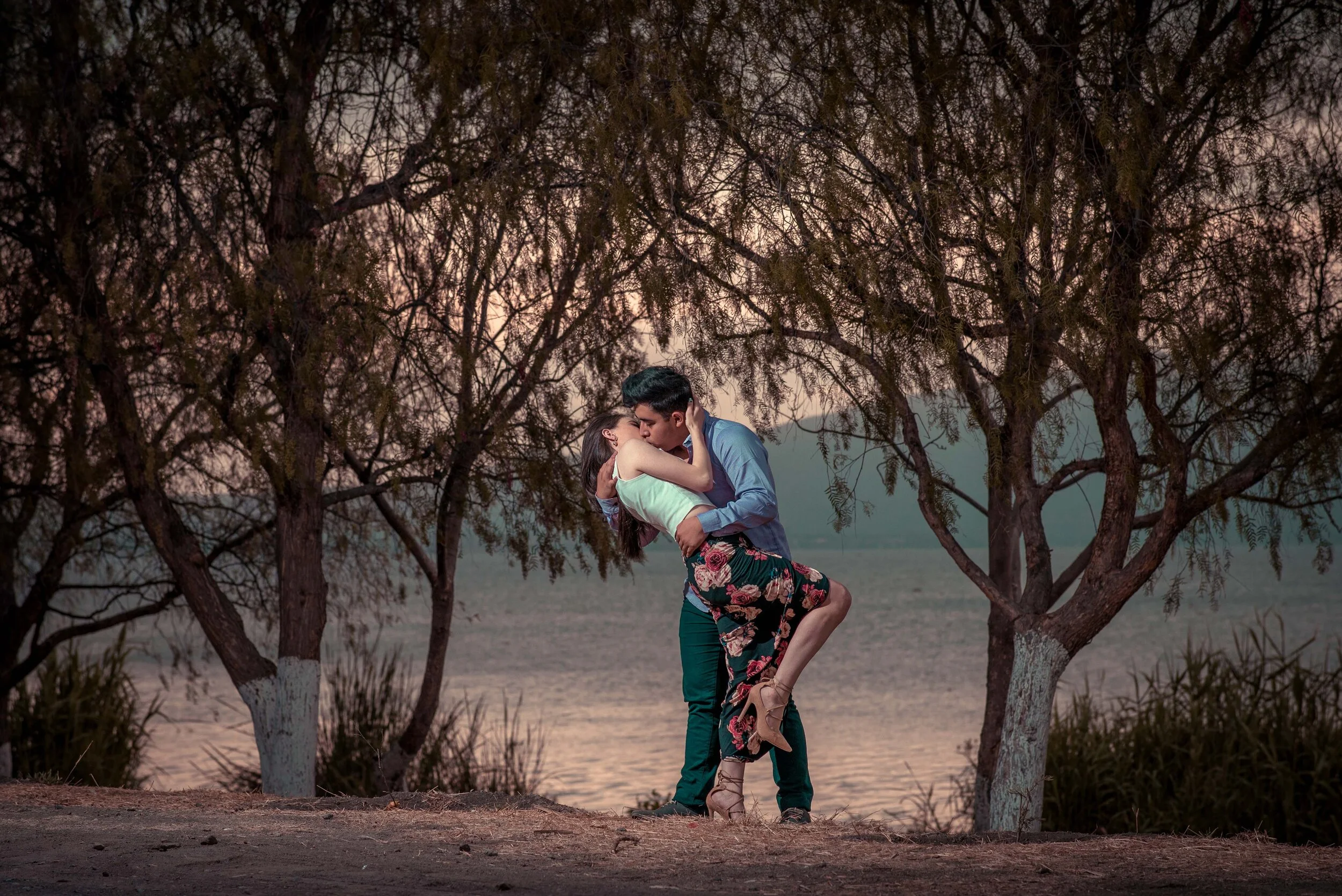 A couple kissing under trees by a lake at sunset.