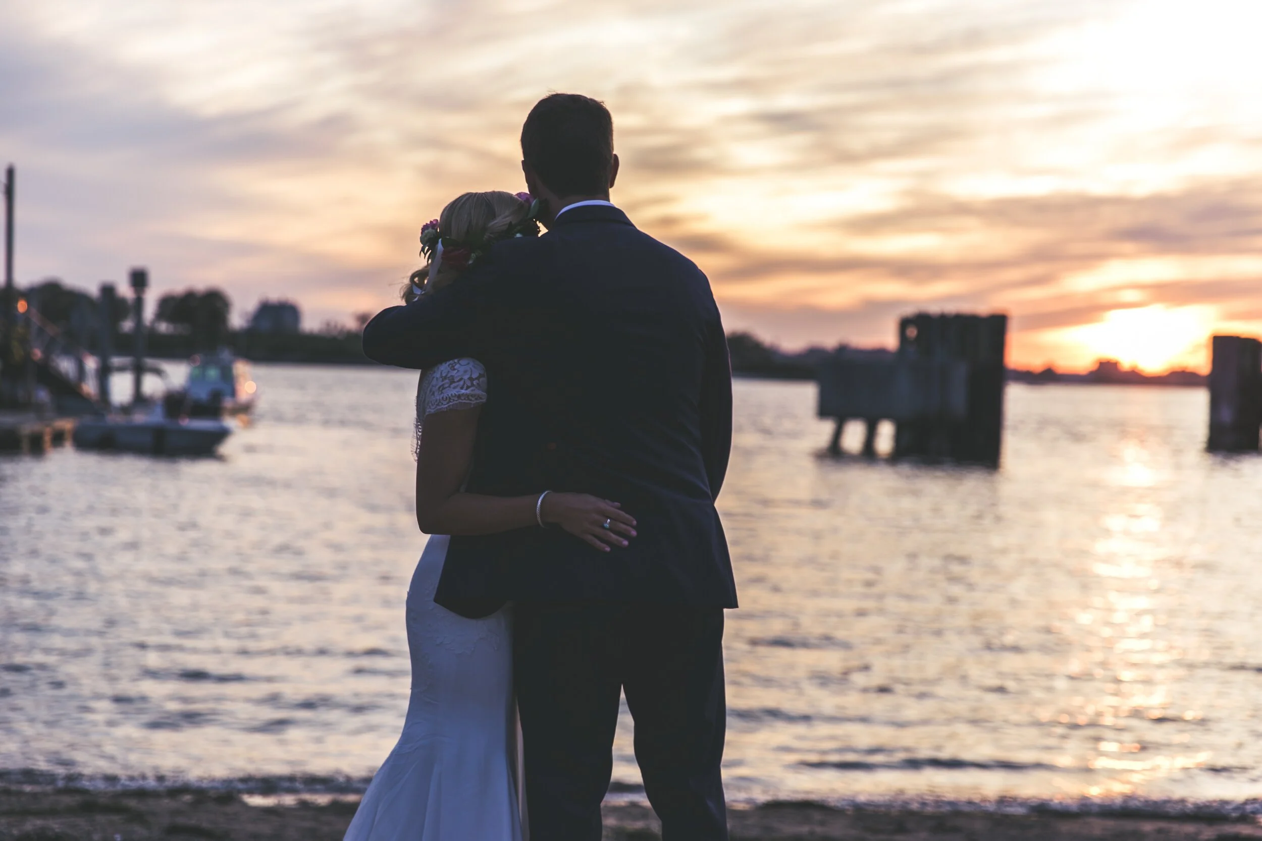 Couple embracing by the waterfront at sunset, with a dock and boats in the background.