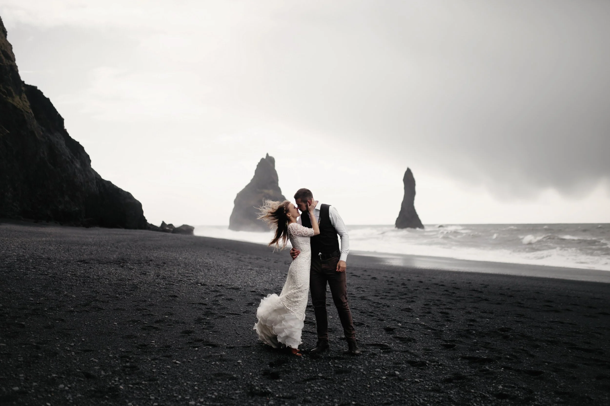 Couple kissing on black sand beach with sea stacks in background, overcast sky.