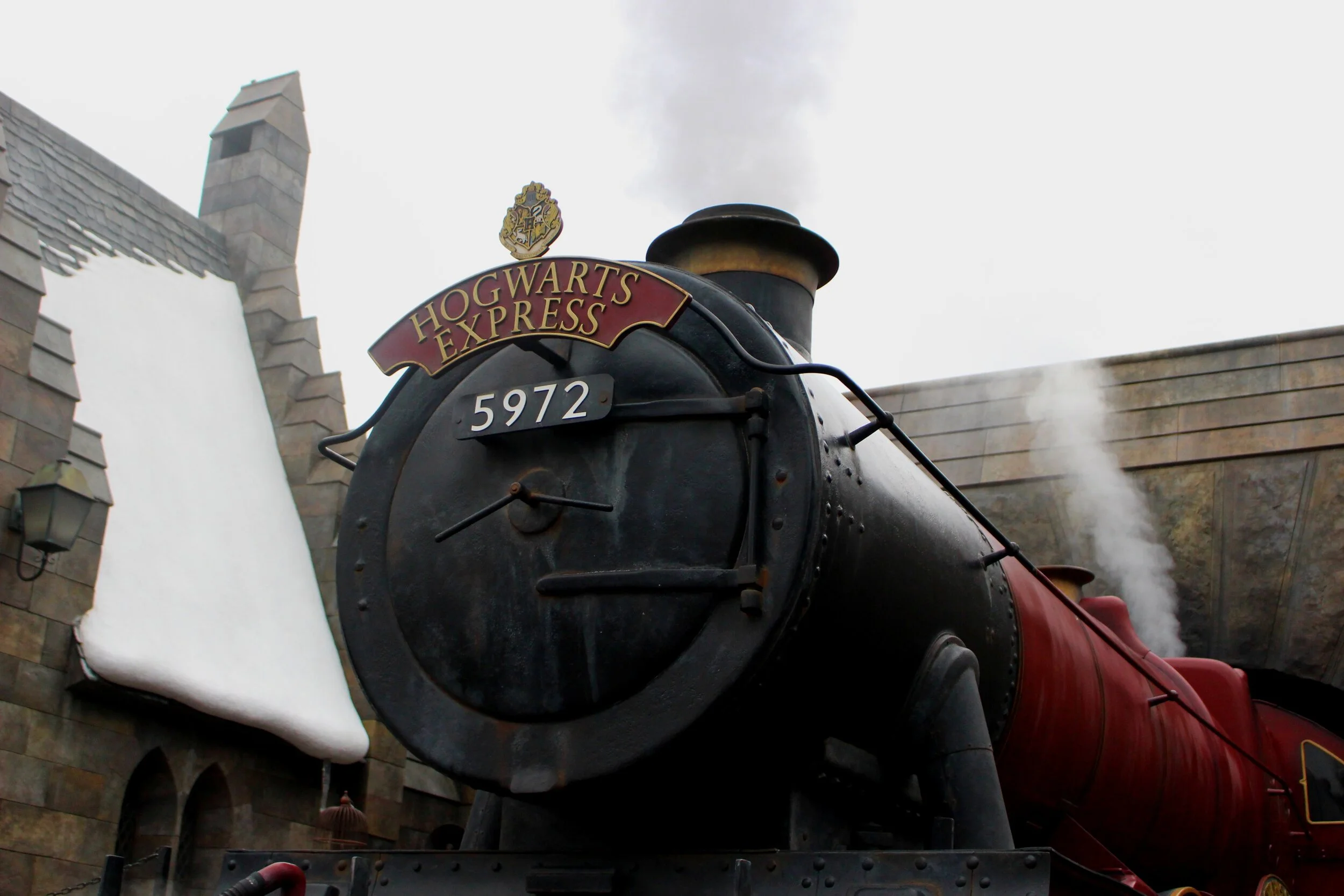 Hogwarts Express train with 5972 written on the front, steaming, and snow-covered building in the background.