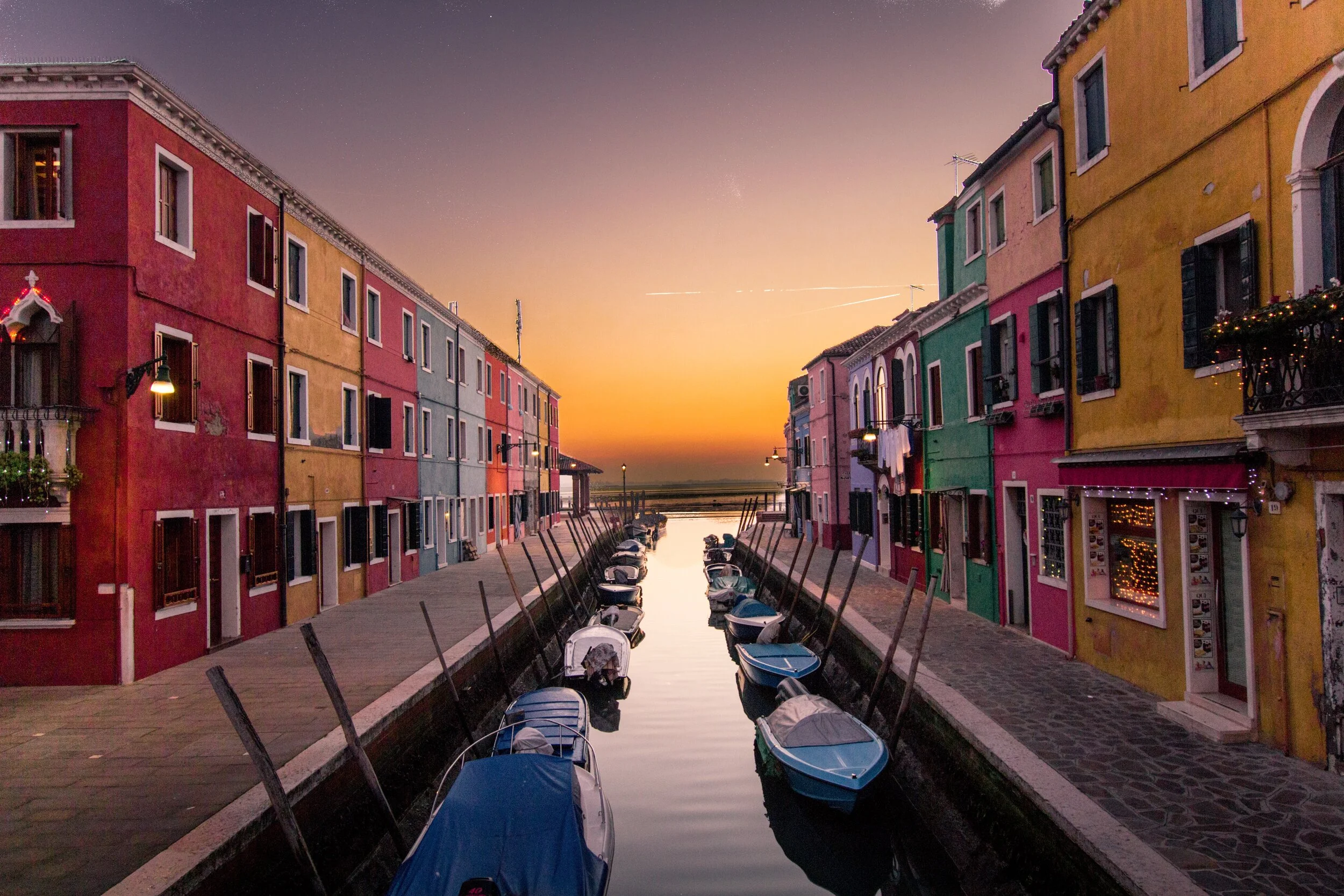 Colorful houses lining a canal in Burano, Italy, with boats on the water at sunset.