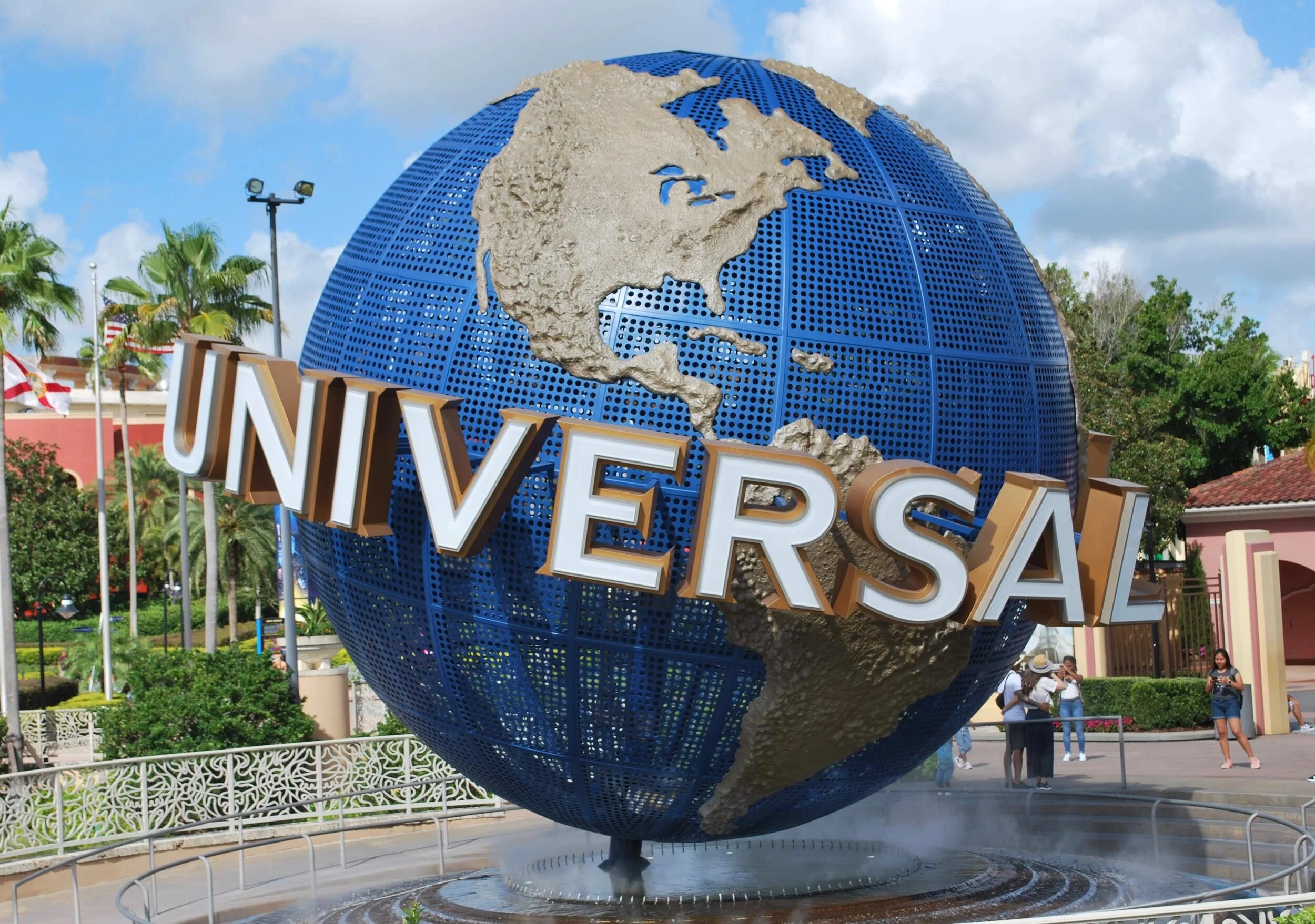 Universal Studios globe sign at an entrance with people around, palm trees, and a blue sky.