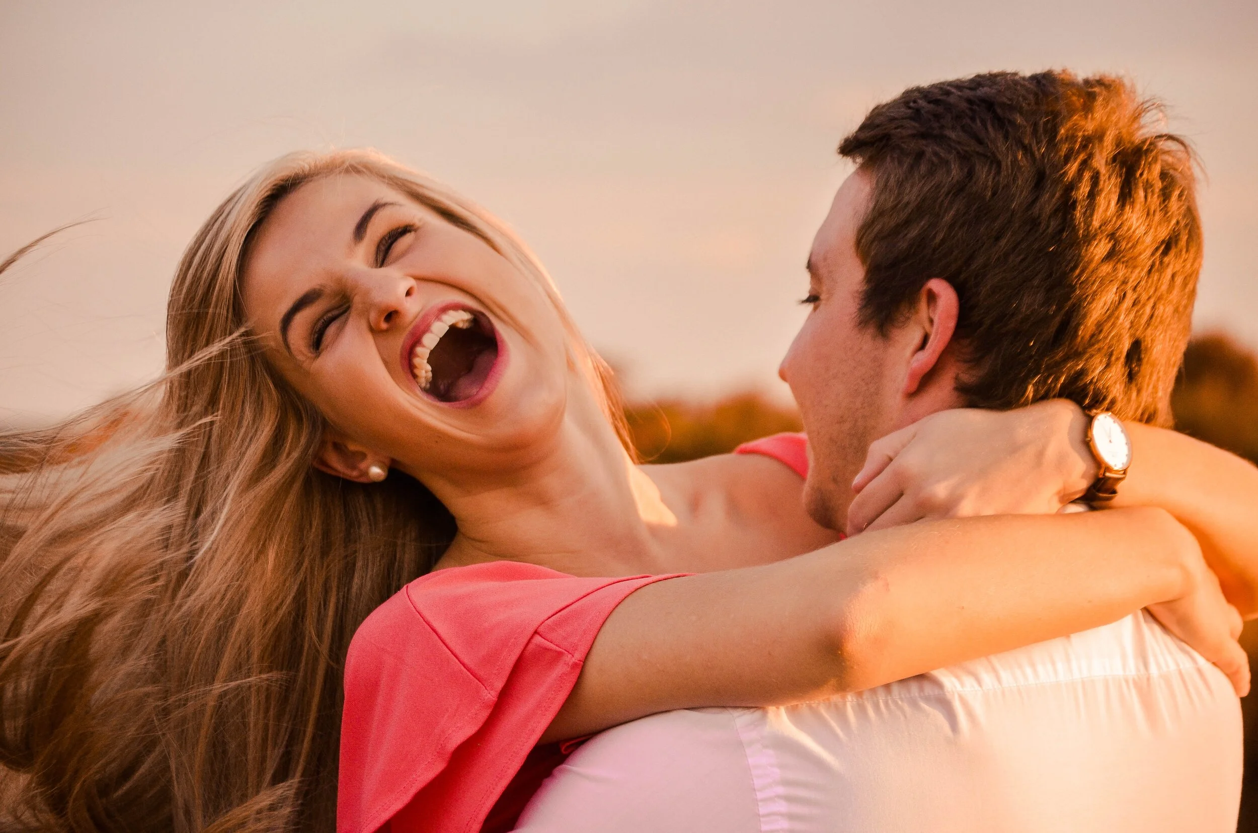 A joyful couple embracing, with the woman laughing heartily while being held by a man, set against a sunset background.