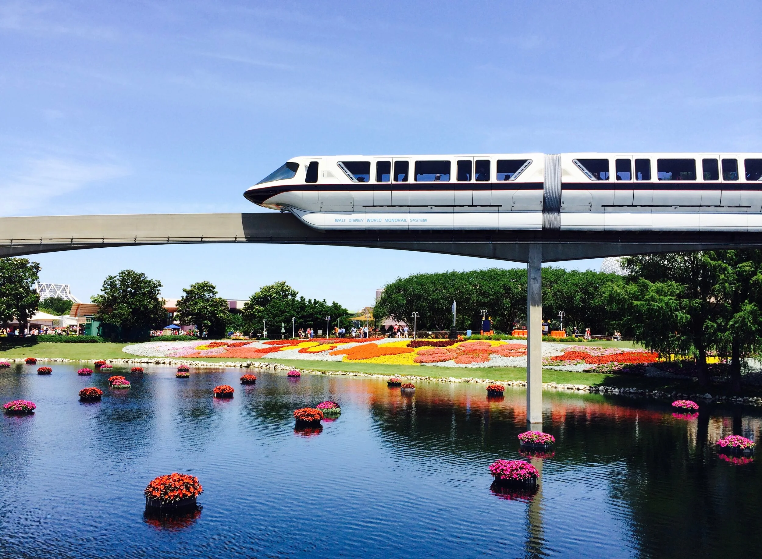 Monorail passing over a lake with floating flower arrangements, vibrant flower beds, and trees in the background.