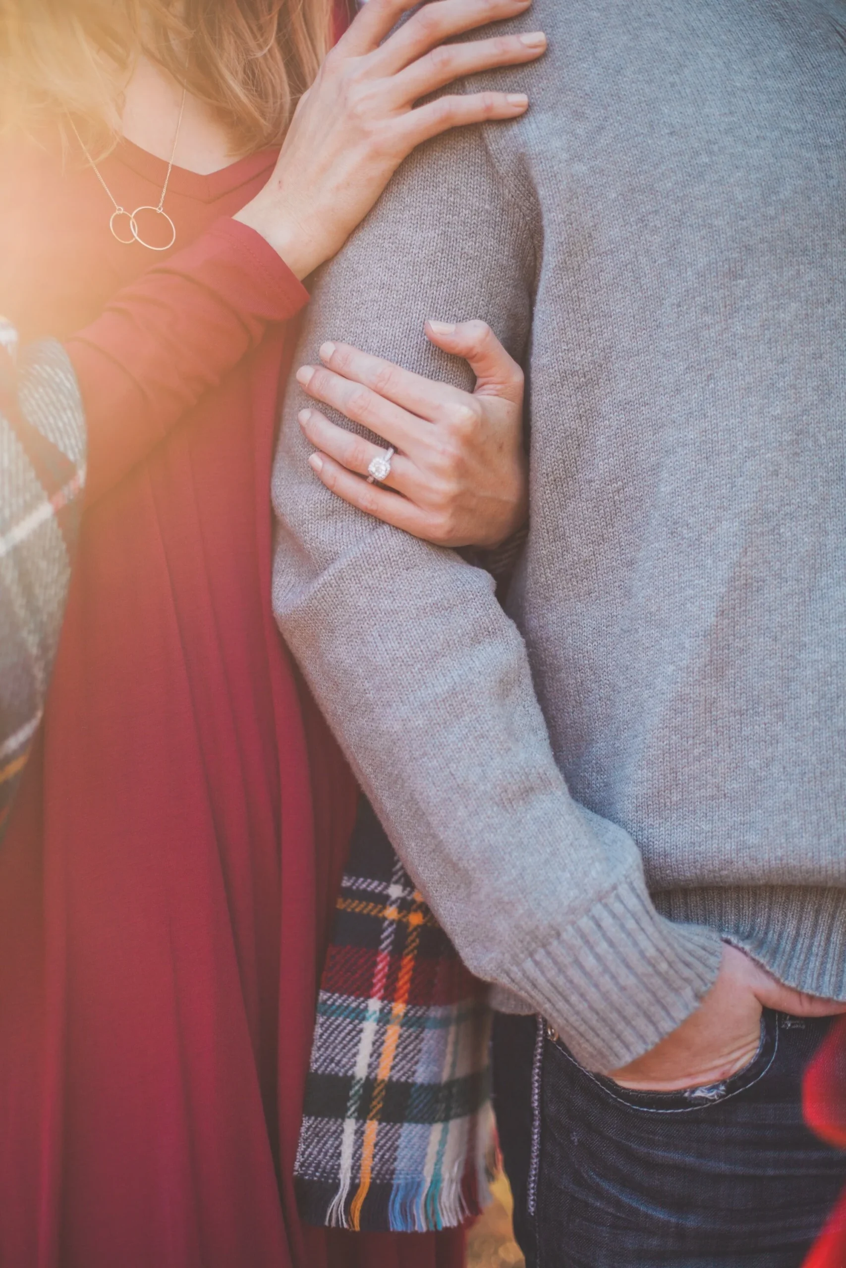 Close-up of a couple embracing, wearing a red dress and a gray sweater. The woman shows a diamond ring on her finger and has her arm around the man's arm, who has his hand in his pocket. A plaid scarf is visible.