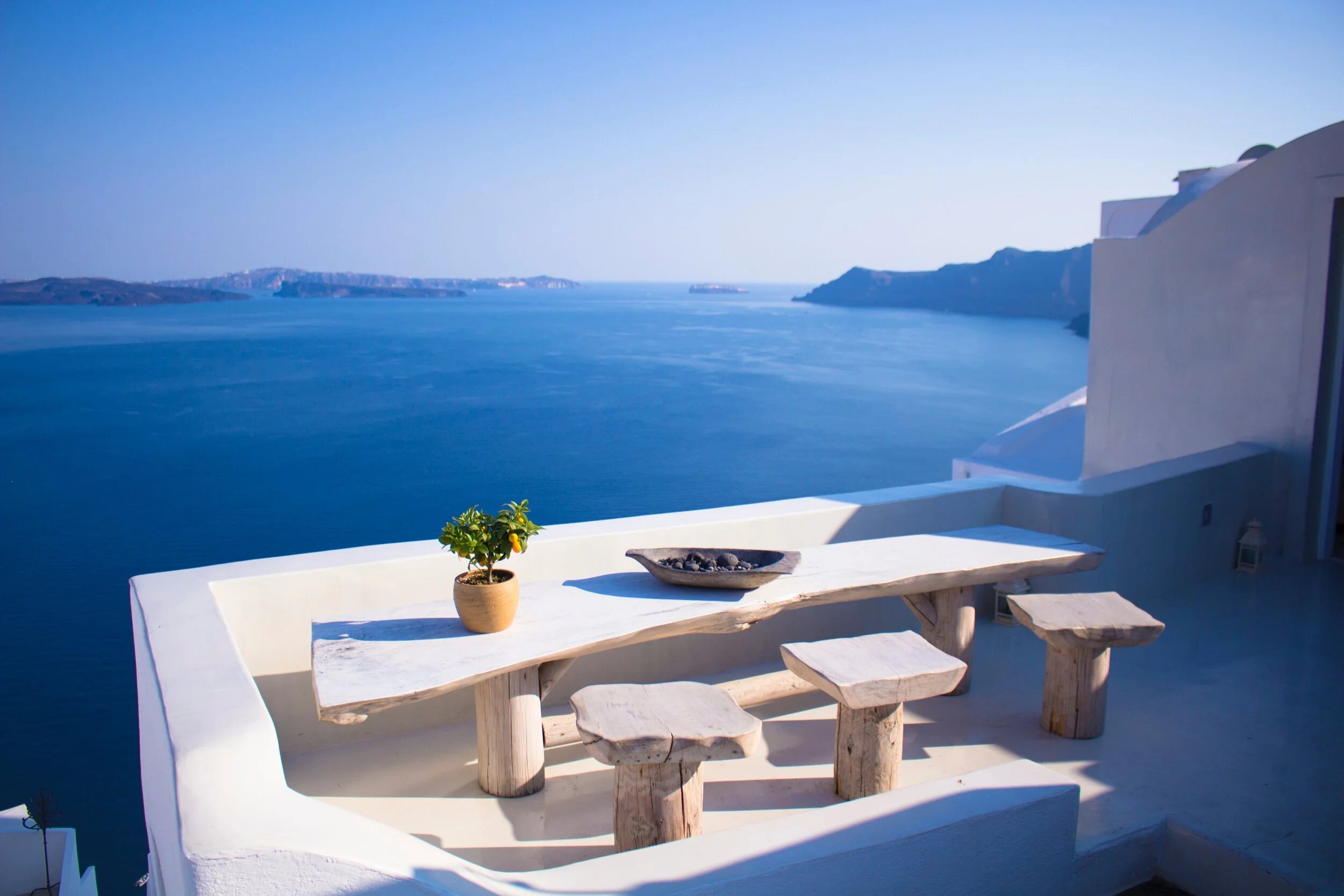 Outdoor terrace overlooking the sea with a wooden table and stools, decorated with a potted plant and bowl, against a clear blue sky.