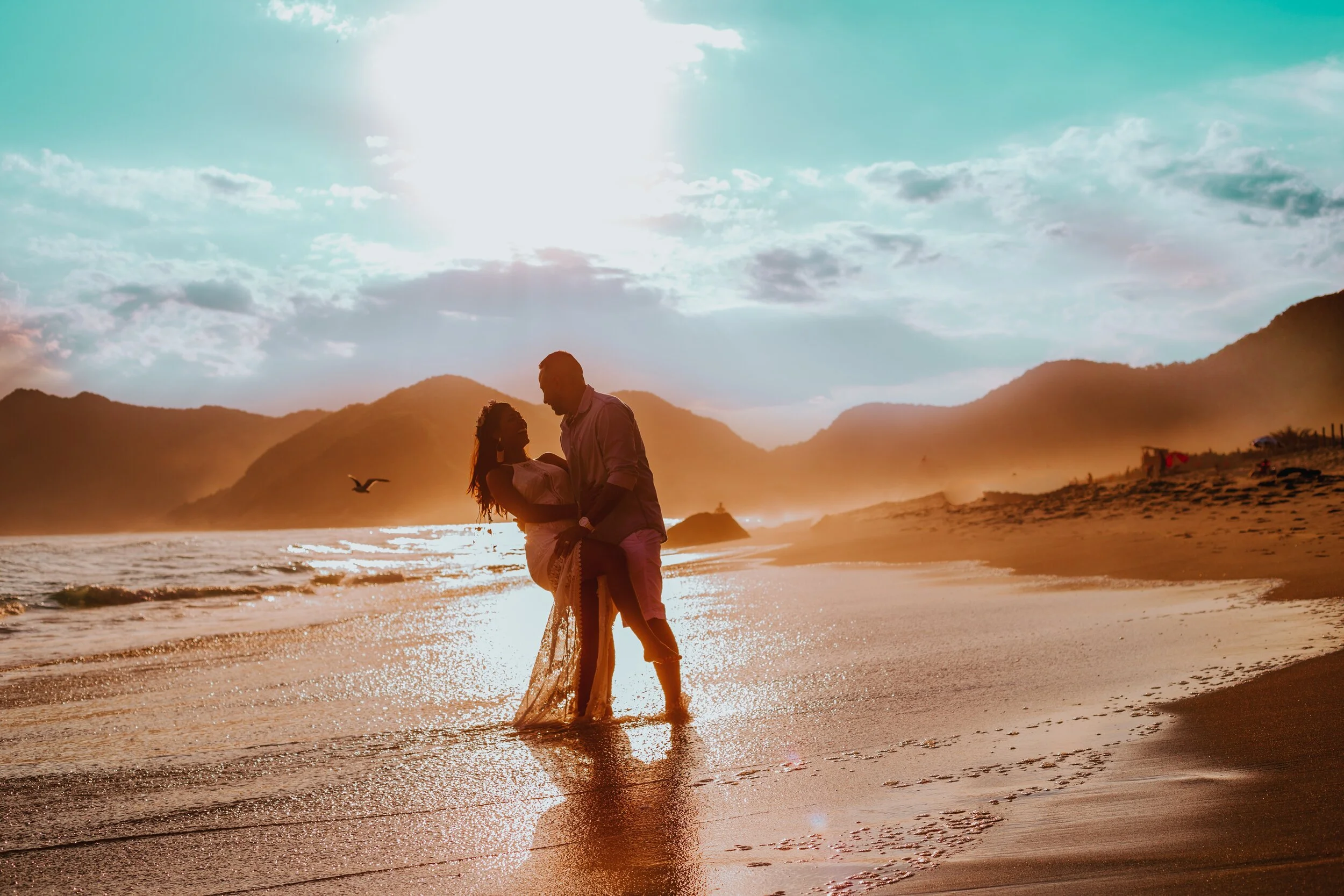Couple embracing on a beach at sunset with mountains in the background.