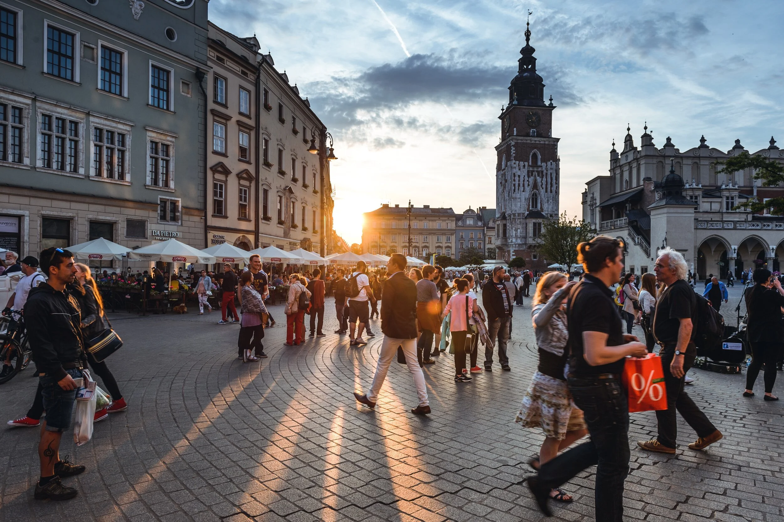 People walking in a bustling European city square at sunset, with historic buildings and a clock tower in the background.