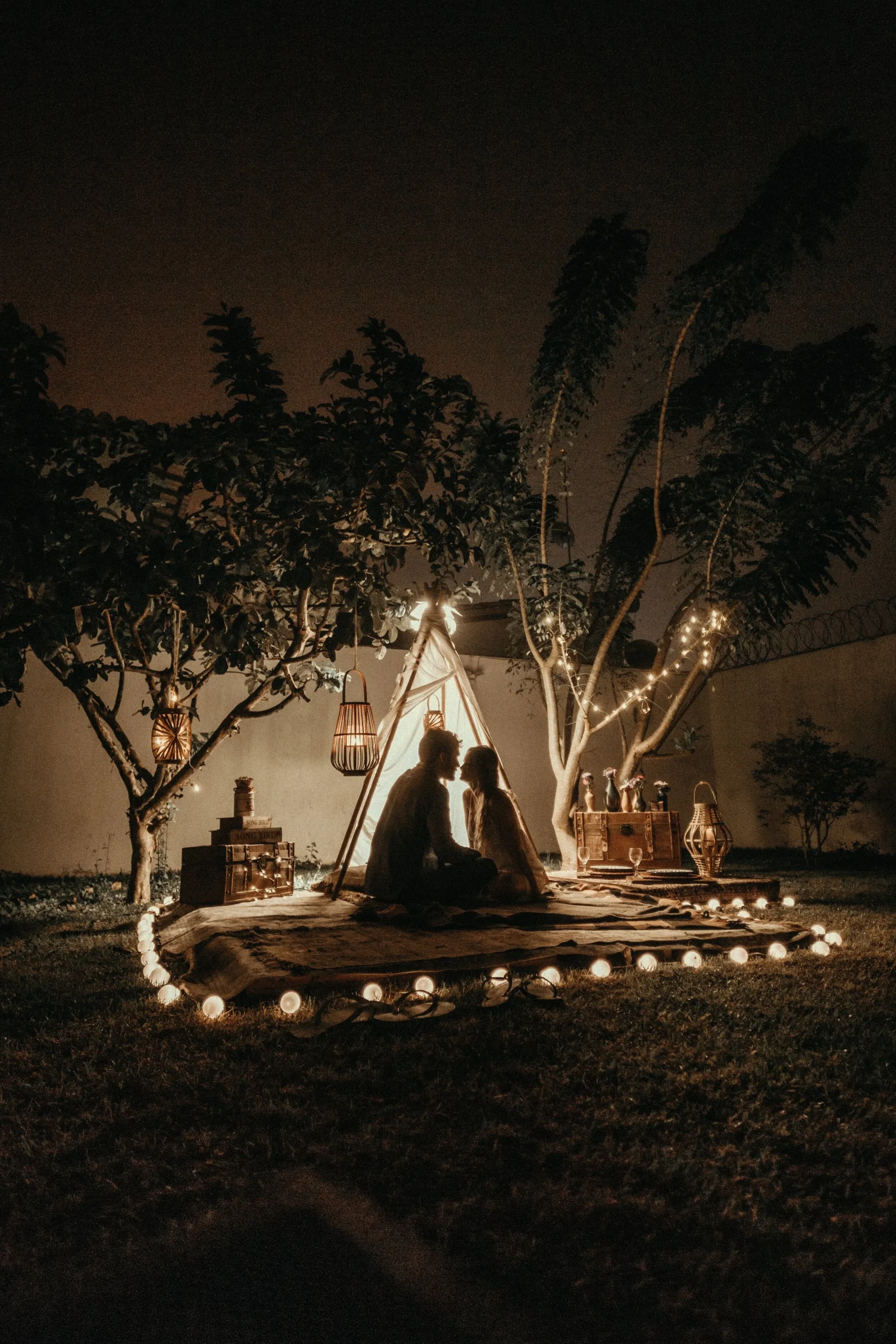 A romantic outdoor evening scene with a couple sitting near a small lit-up tent, surrounded by hanging lanterns and string lights in a garden setting.