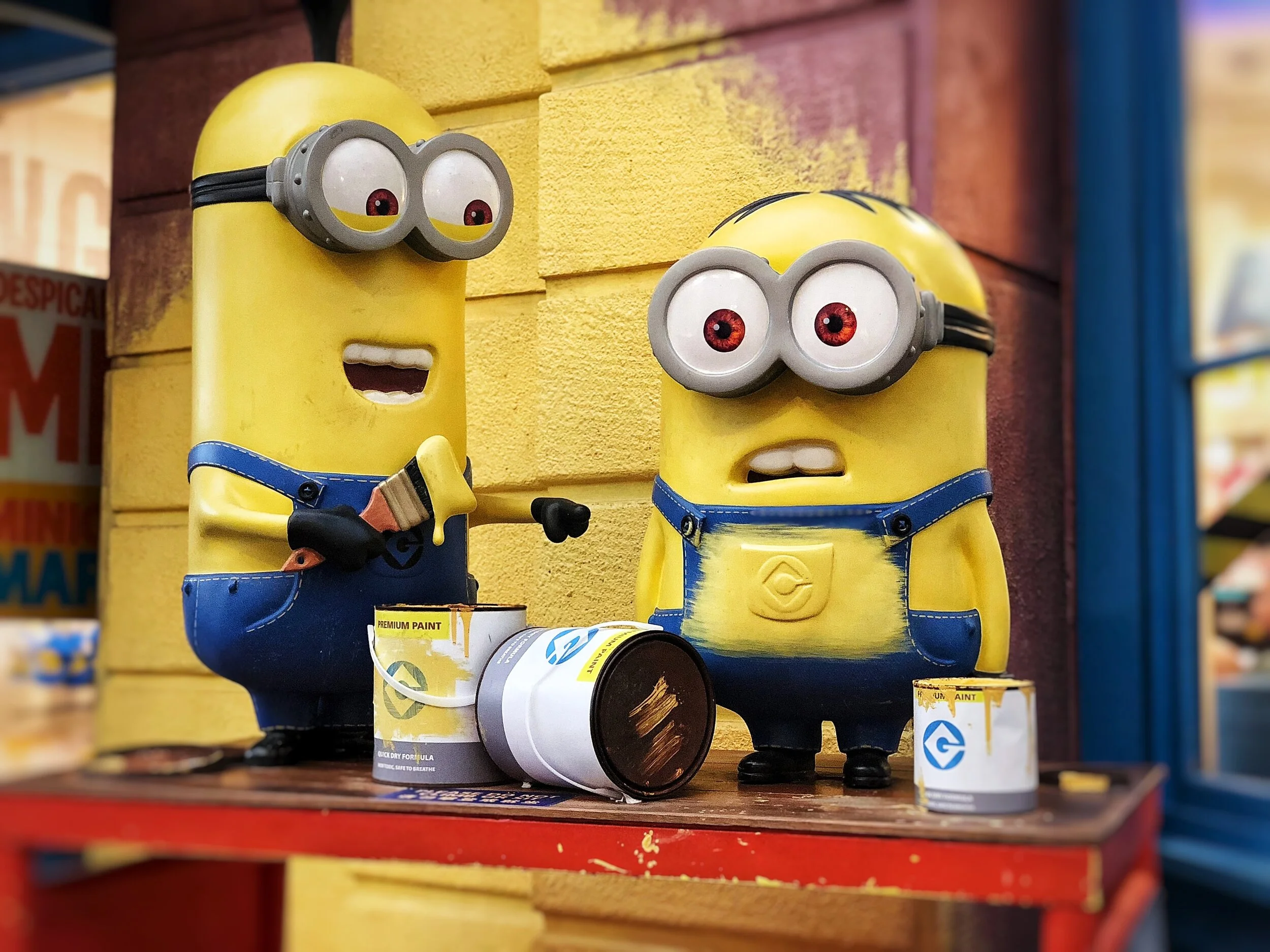 Two cartoon Minion characters with paint cans, one holding a paintbrush, against a yellow wall backdrop.