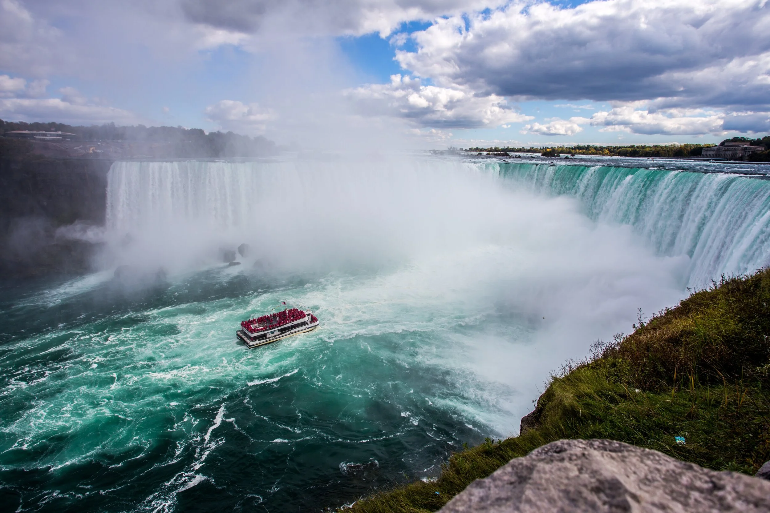 Tour boat at Niagara Falls with mist and cascading water, surrounded by greenery and a cloudy sky.