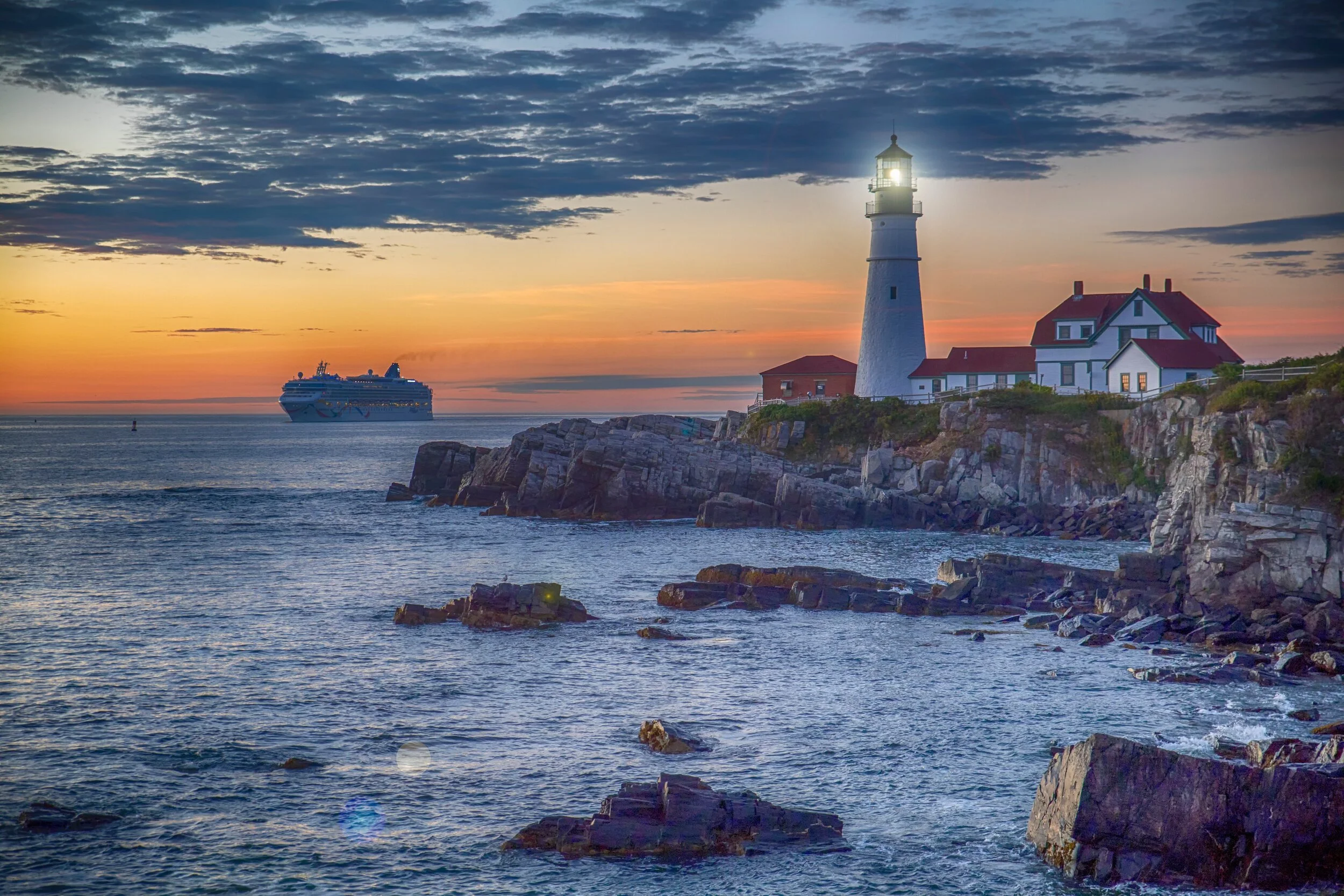 Lighthouse on rocky coast at sunset with a cruise ship in the ocean.