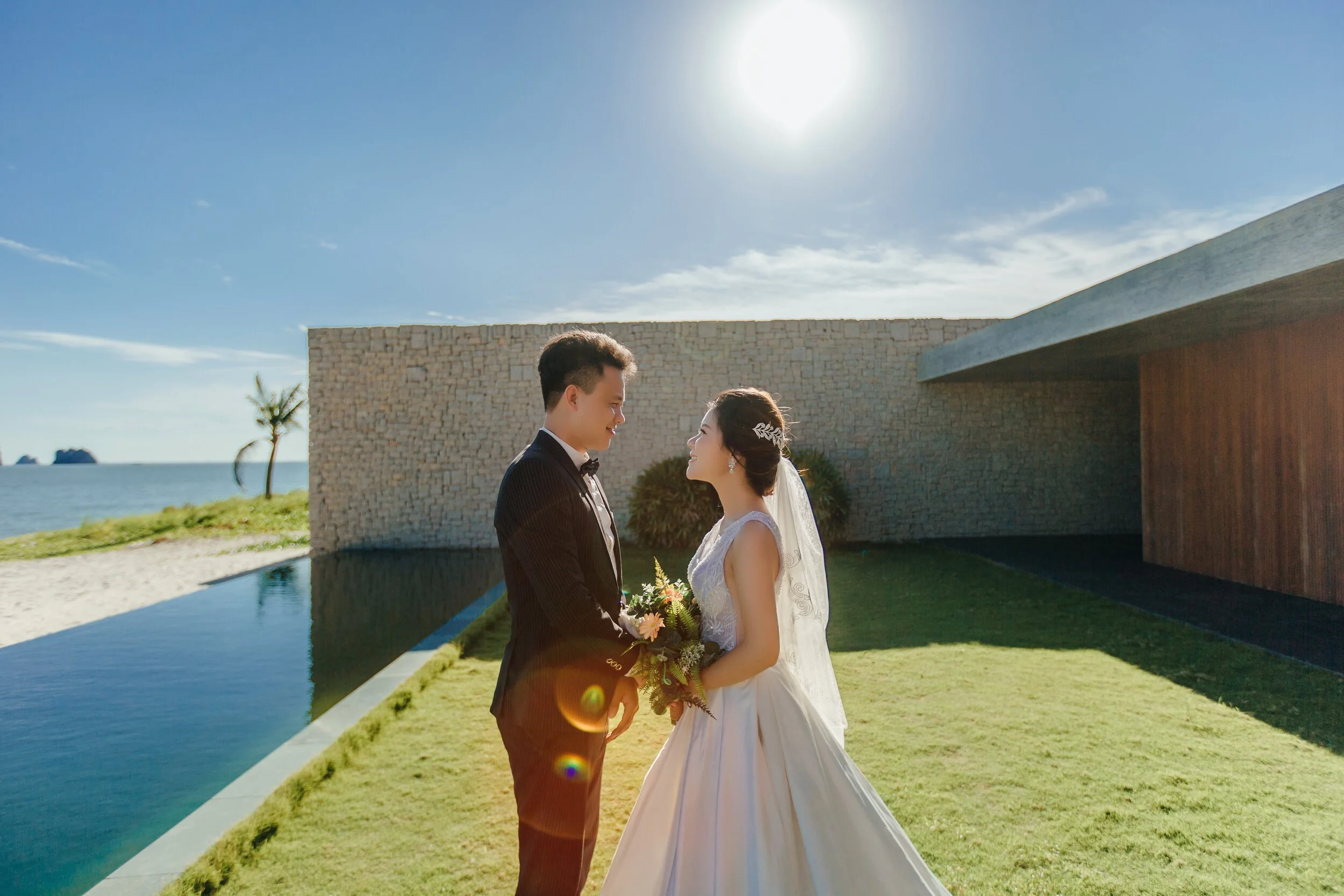 A bride and groom facing each other outdoors by a modern building with a stone wall, near a body of water and a small palm tree, under a sunny sky.
