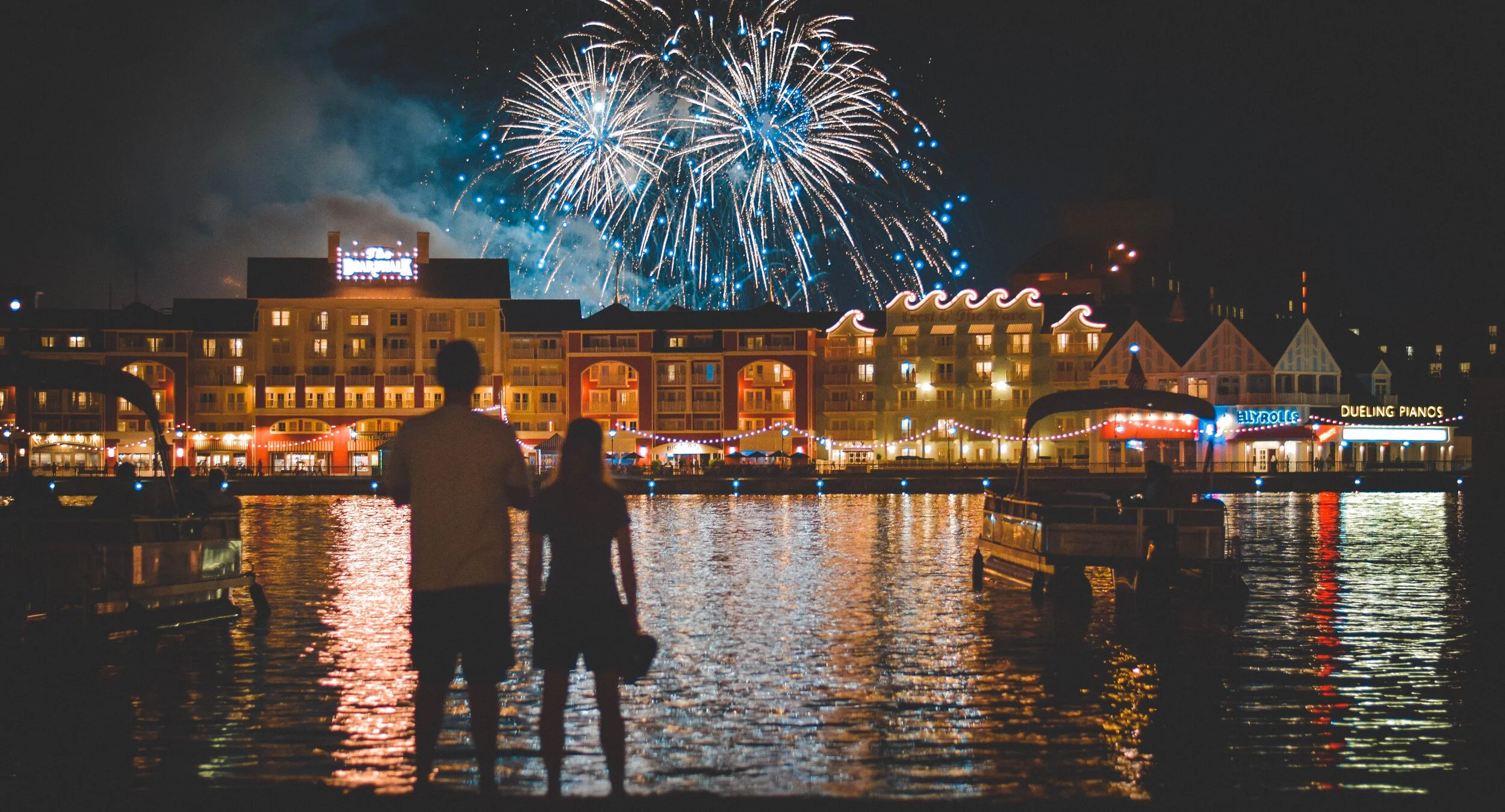 Silhouetted couple watching fireworks over a waterfront with buildings and boats at night.