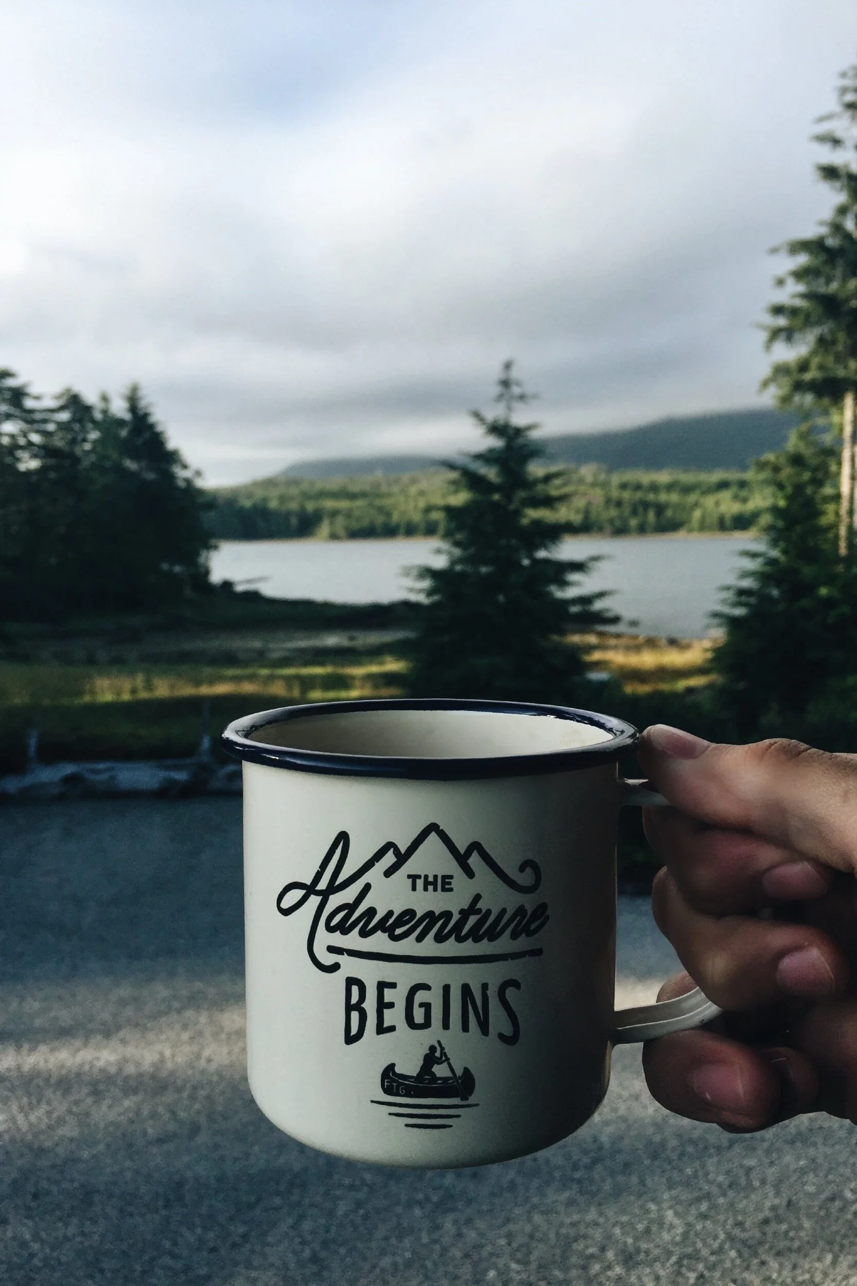 Person holding a mug with "The Adventure Begins" text, in front of a scenic view with trees, a lake, and a cloudy sky in the background.