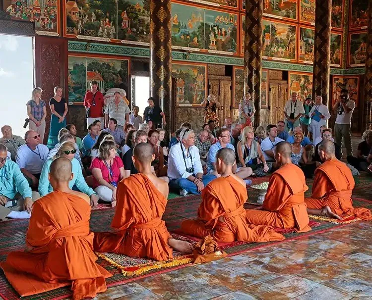 Group of monks in orange robes sitting in a temple with an audience, surrounded by colorful murals and ornate architectural details.