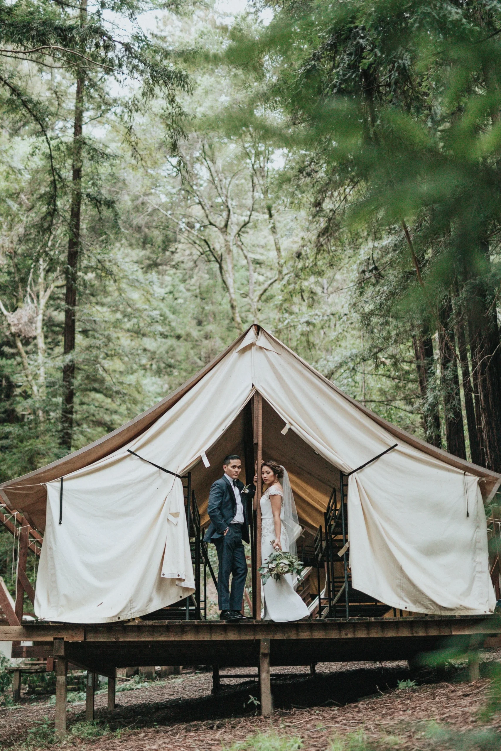 Bride and groom standing in front of a glamping tent in a forest setting.