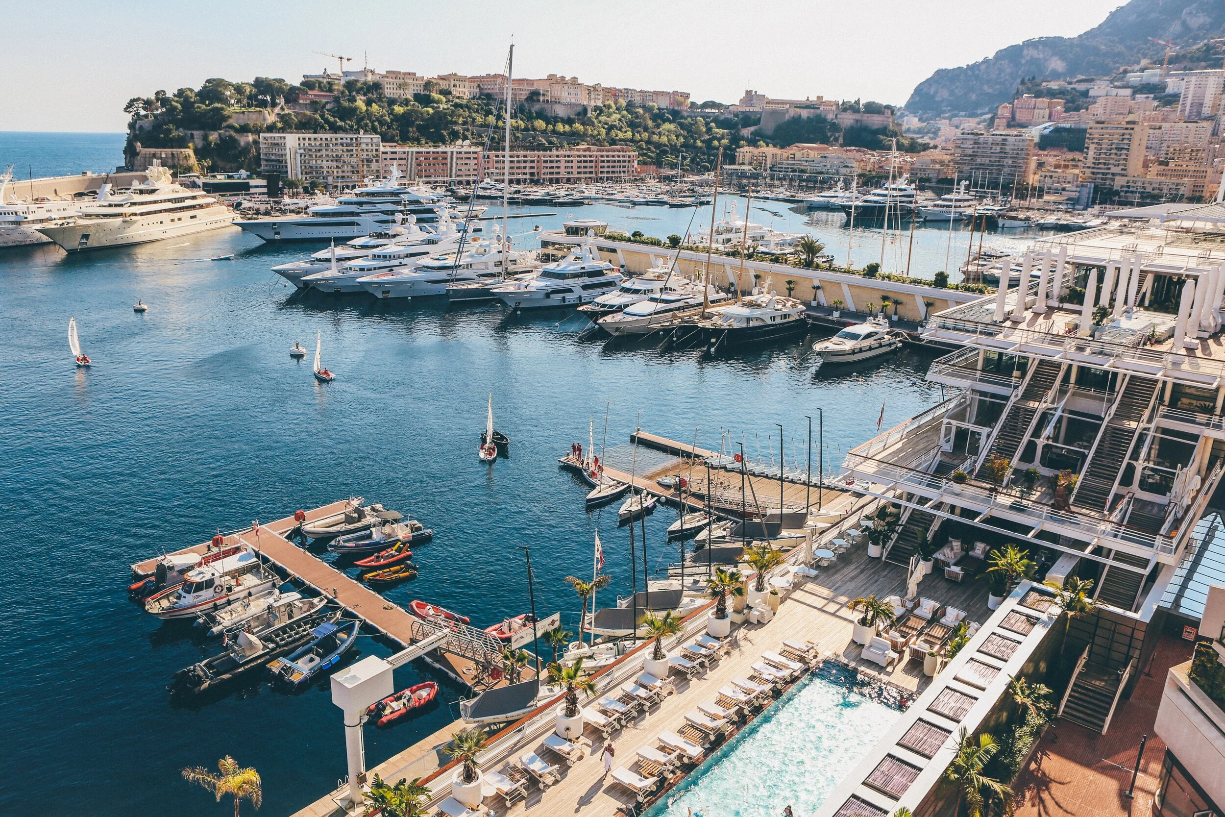 View of a luxurious marina with yachts docked in the harbor, surrounded by a hilly cityscape and waterfront buildings. The scene includes small boats sailing in the water and a foreground area with a pool, deck chairs, and palm trees, emphasizing a l