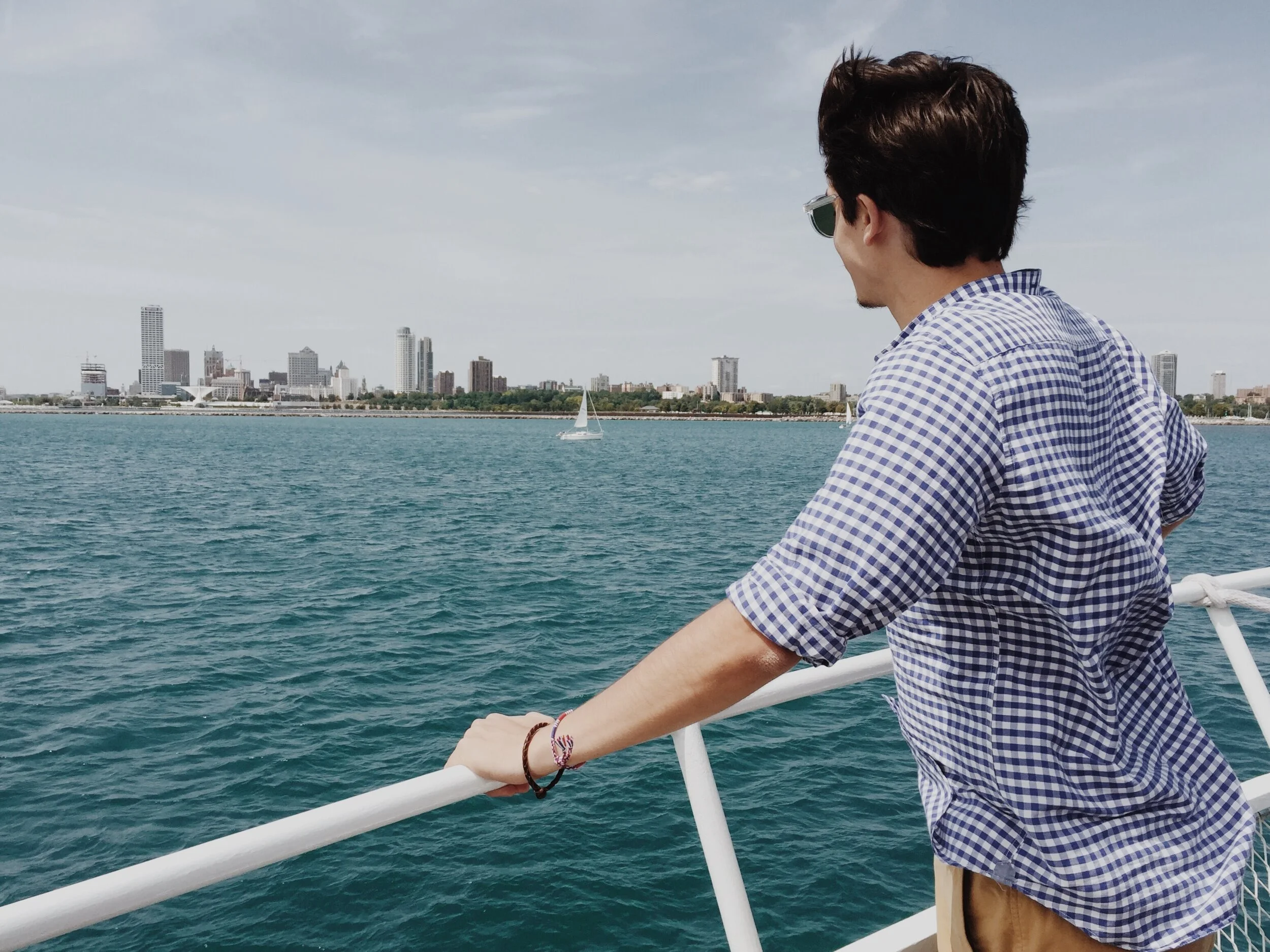Person in a checkered shirt on a boat, overlooking a city skyline across a body of water, with a sailboat in the distance.