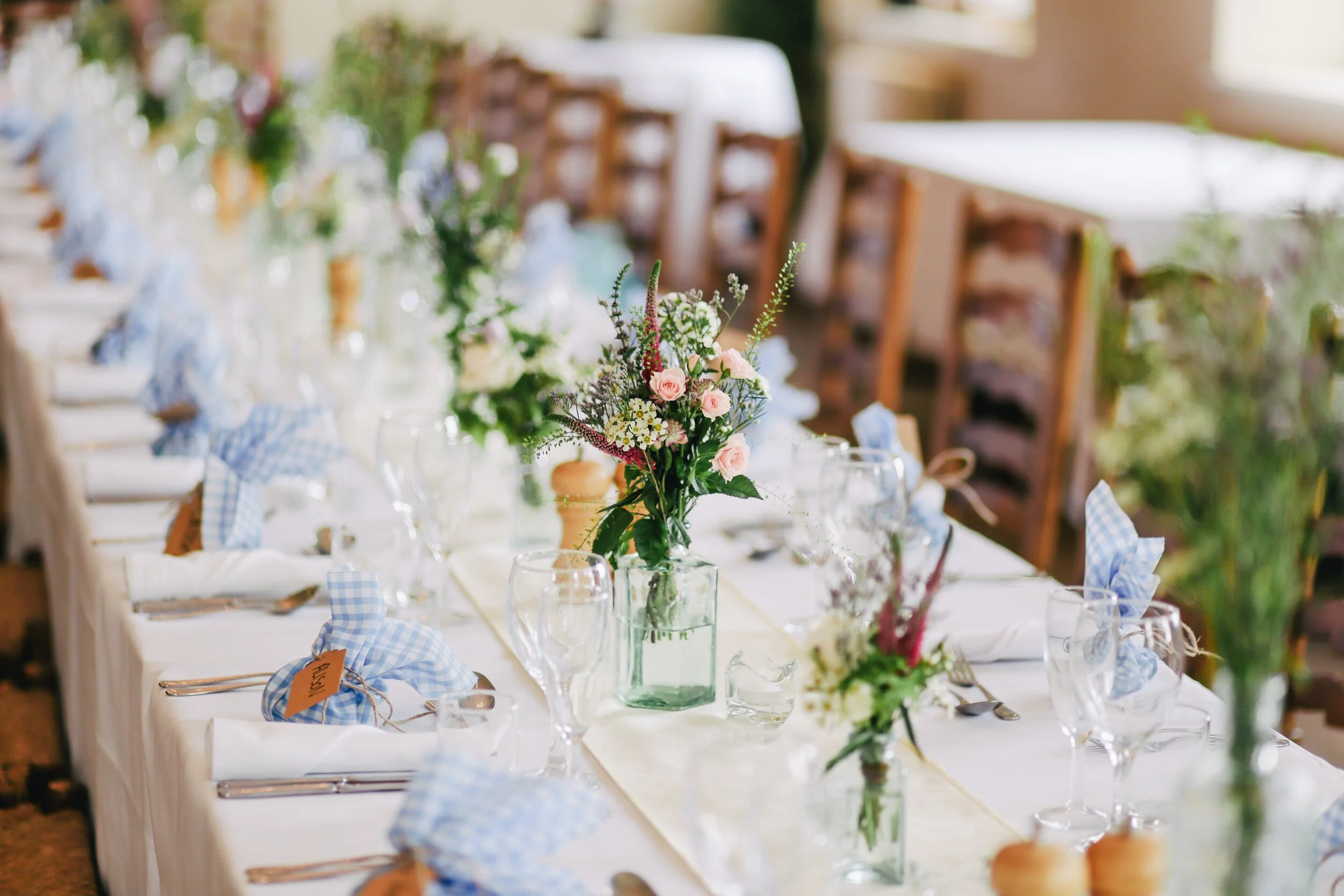 Elegant table setting with white linens, glassware, cutlery, small floral centerpieces, and blue gingham napkins tied with twine on a long dining table.