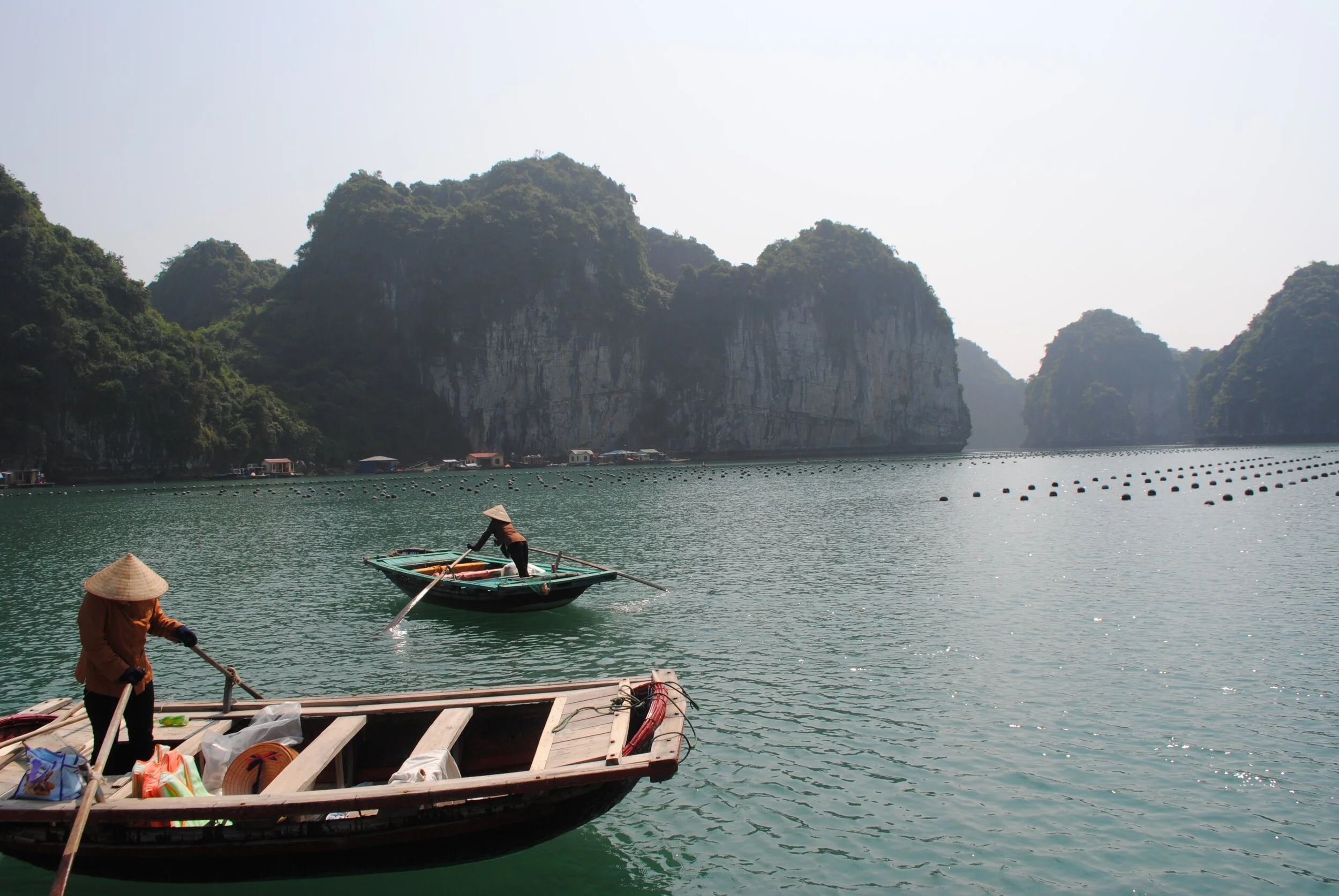 Scenic view of a bay with limestone cliffs, two people on small wooden boats wearing traditional hats, and floating objects on the water, suggesting a fishing or aquaculture area.