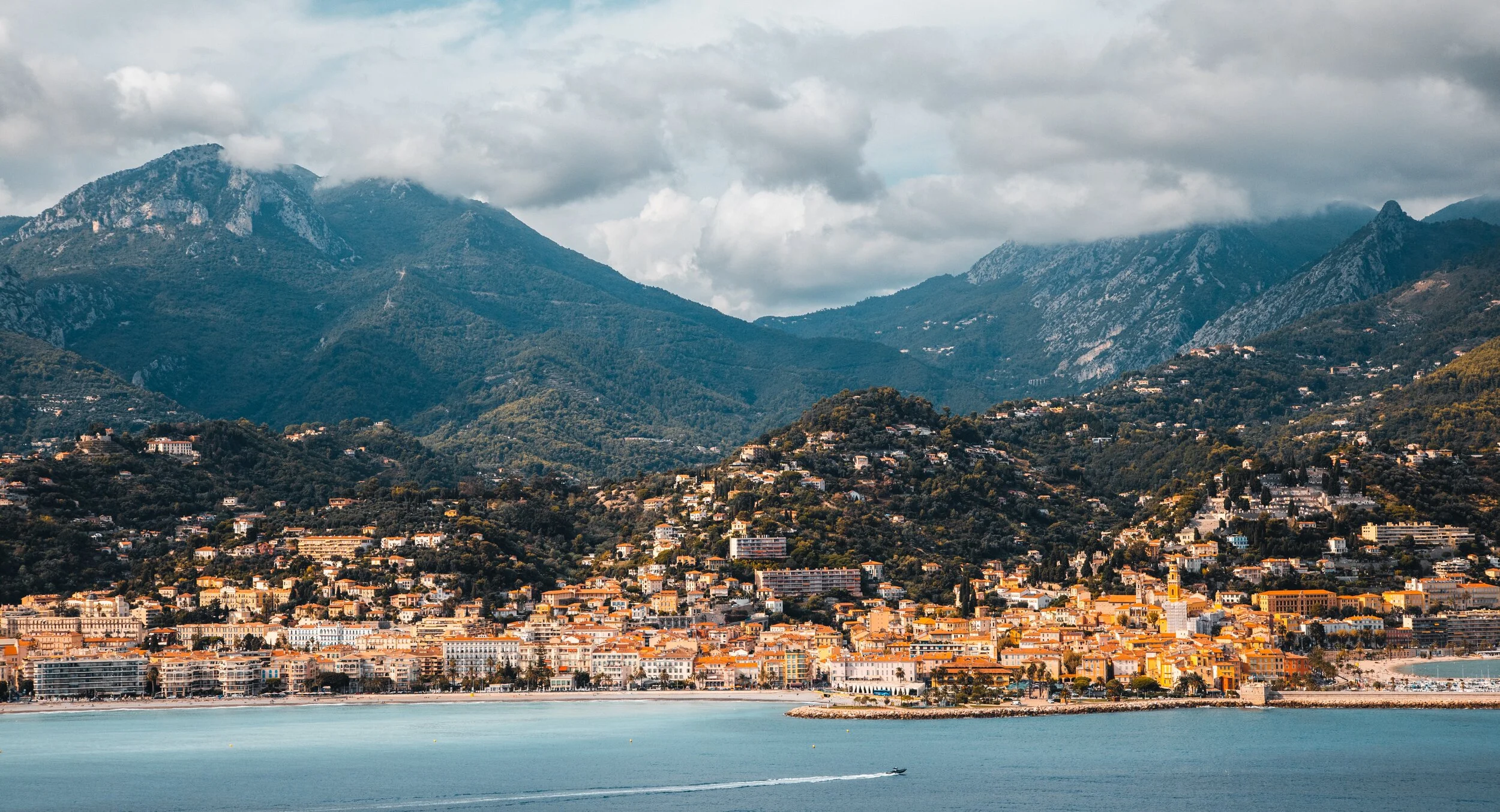 Coastal city with mountains in the background under cloudy sky.