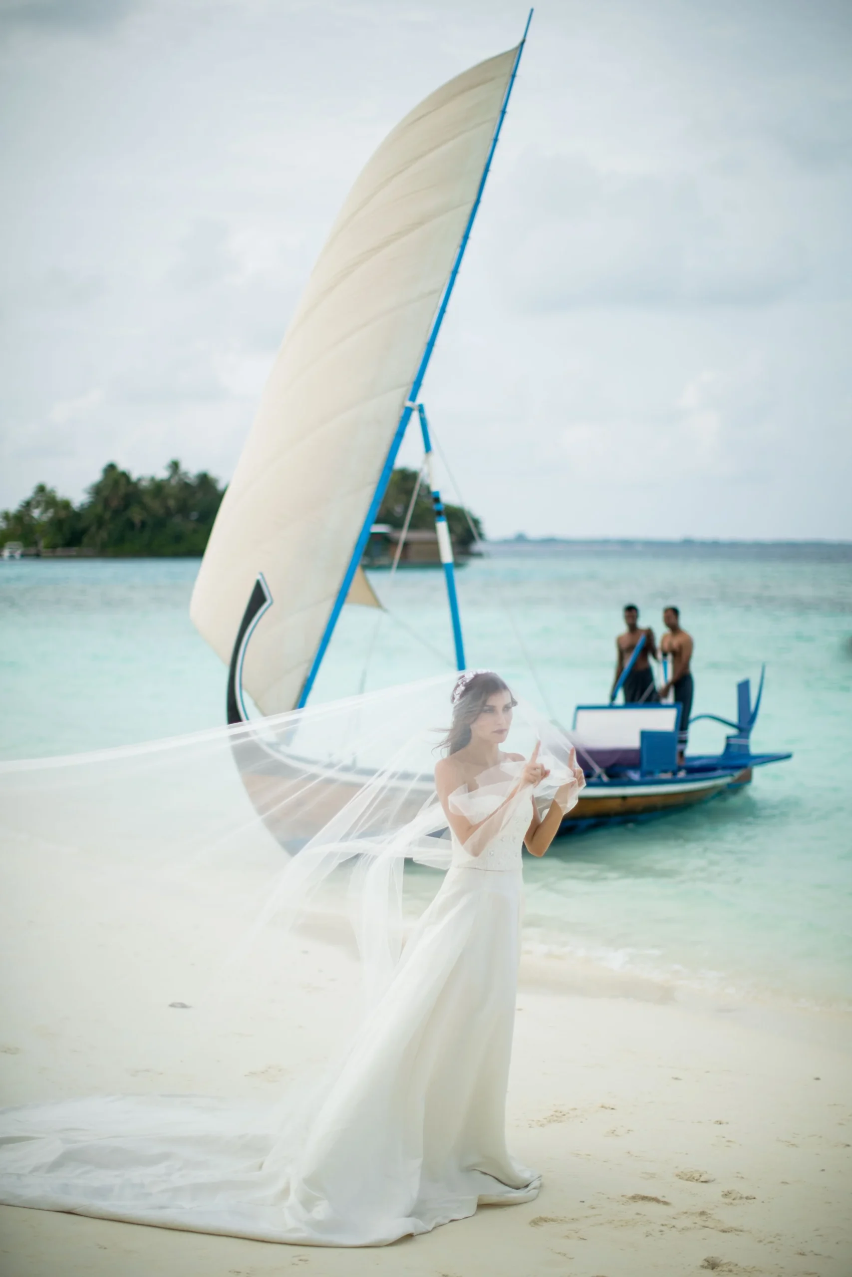 Bride in wedding dress with veil on beach, traditional boat in background, two people onboard.