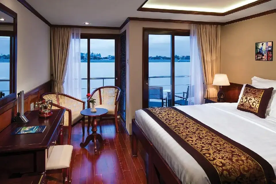 Luxury hotel room with a large bed, elegant wooden furniture, a desk, two chairs around a small table, and a balcony view of water.