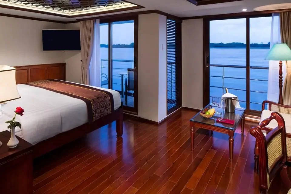 Luxurious bedroom on a cruise ship with wooden flooring, a large bed, elegant decor, glass coffee table with fruit and champagne, and a balcony view of the water.
