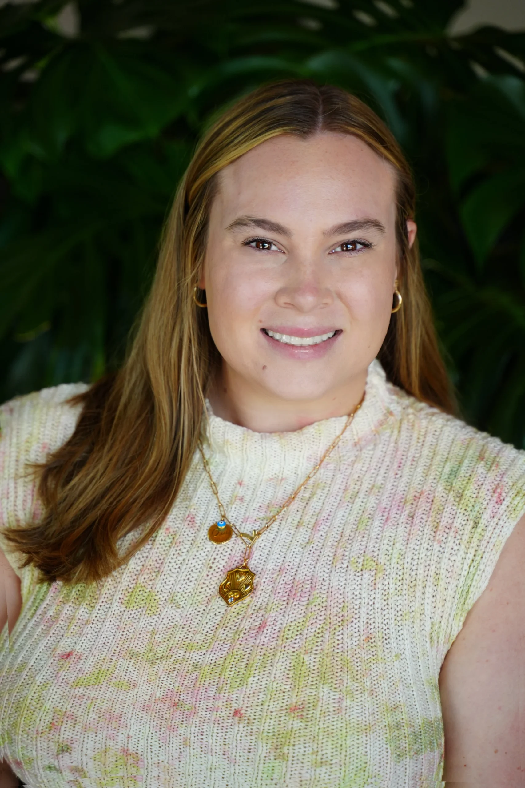 Smiling person wearing a sleeveless knit top and necklace with leafy background.