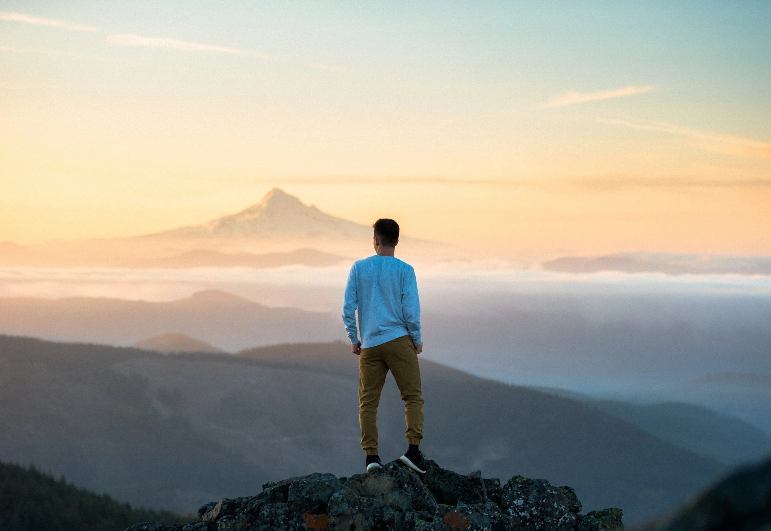 Person standing on a rocky hilltop with a view of distant mountains and a cloudy sky during sunrise or sunset.