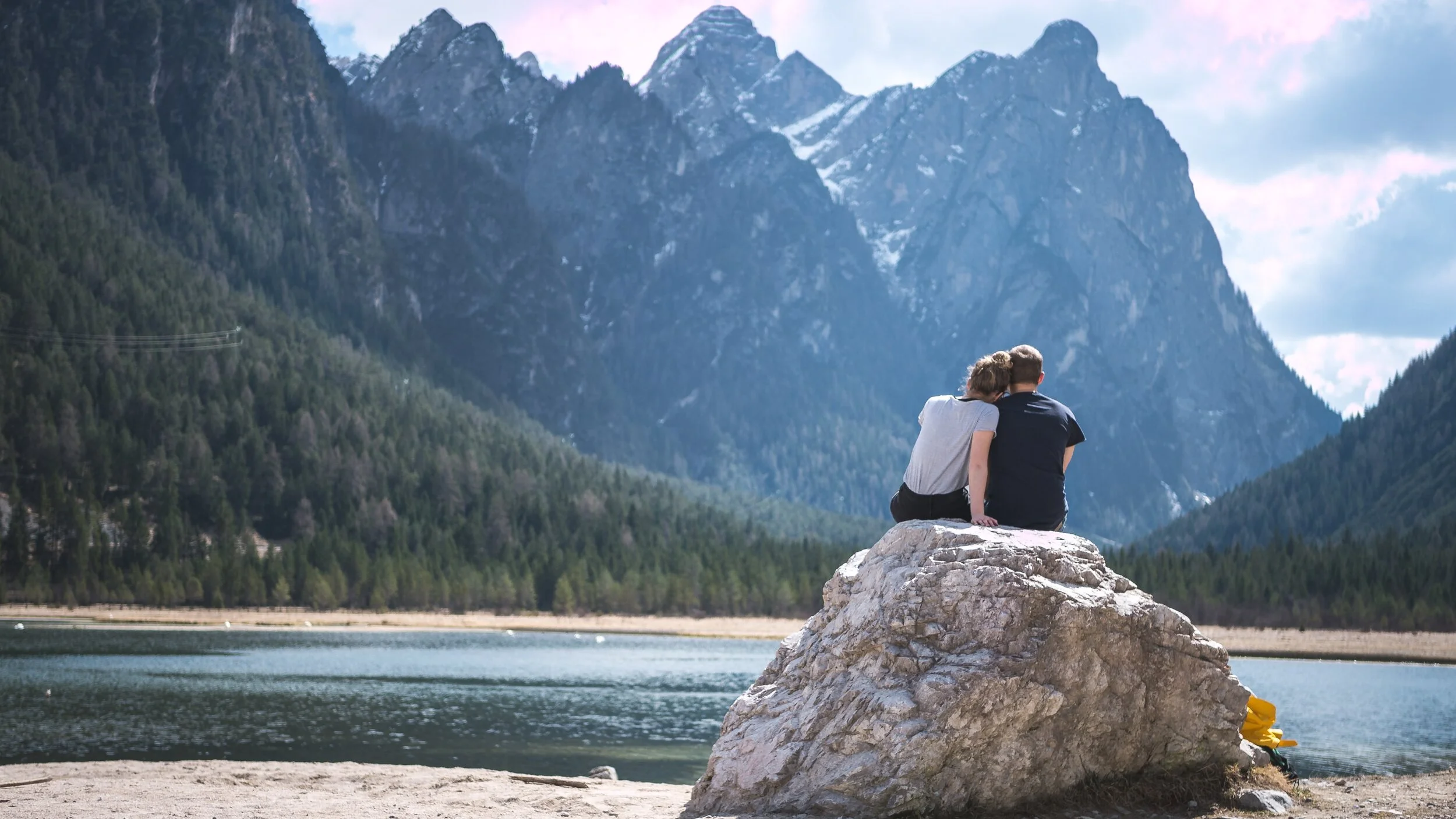 A couple sitting on a large rock by a lake with mountains in the background, enjoying the scenic view.