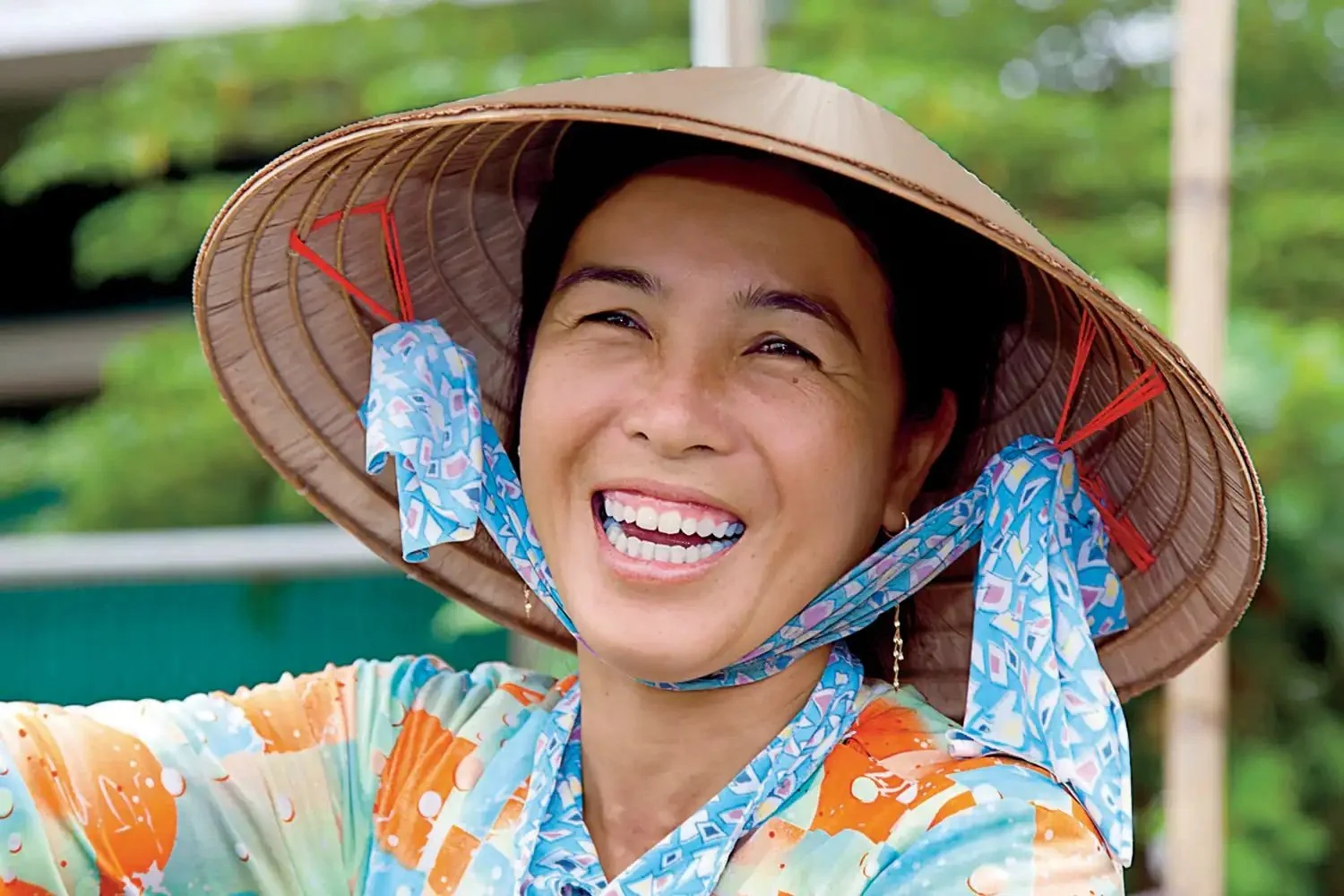 Smiling woman wearing a traditional conical hat and colorful floral clothing outdoors.