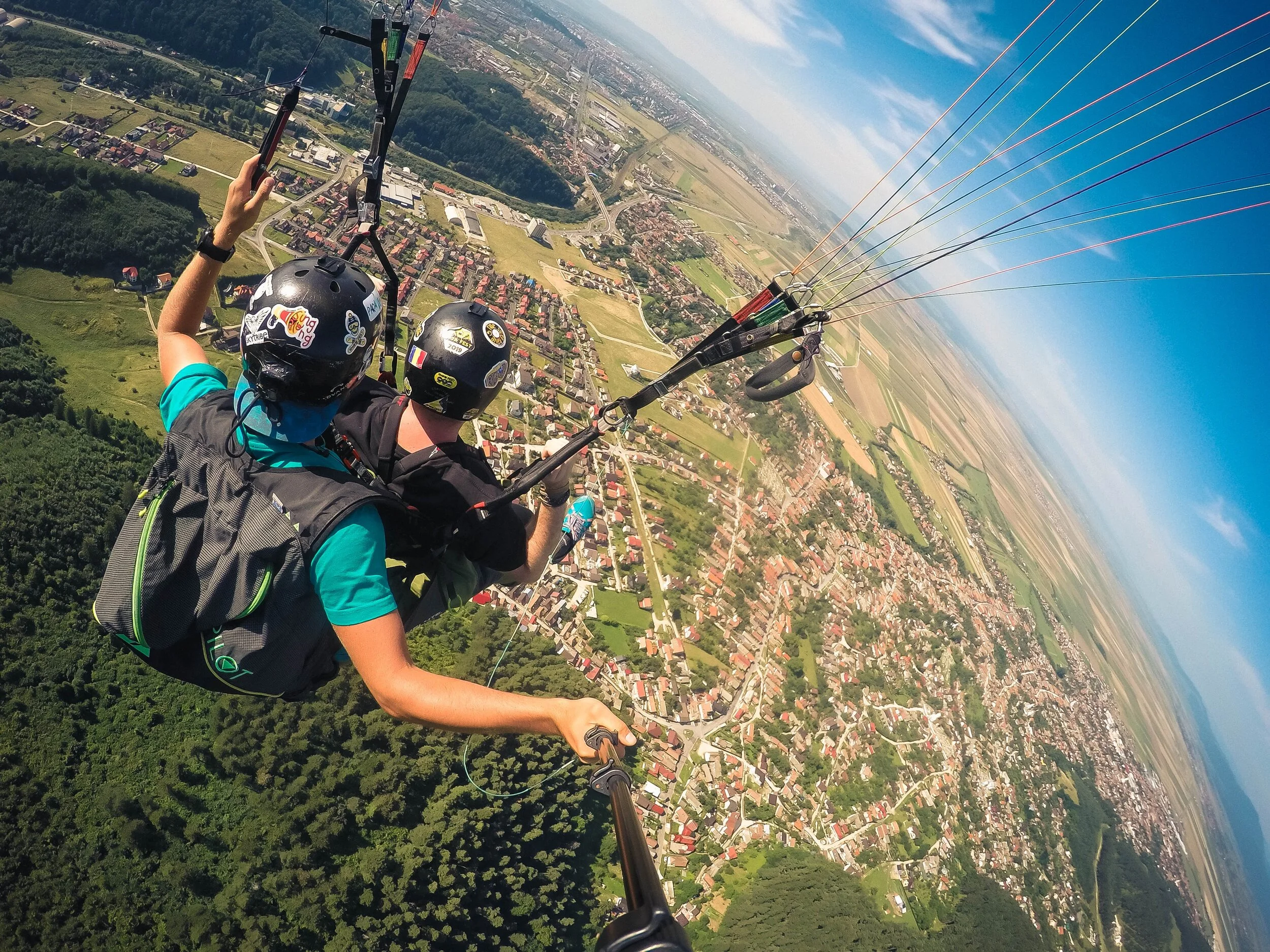 Two people paragliding with a scenic view of a rural landscape, including fields and a small town, beneath them. One person is holding a selfie stick, capturing the moment.